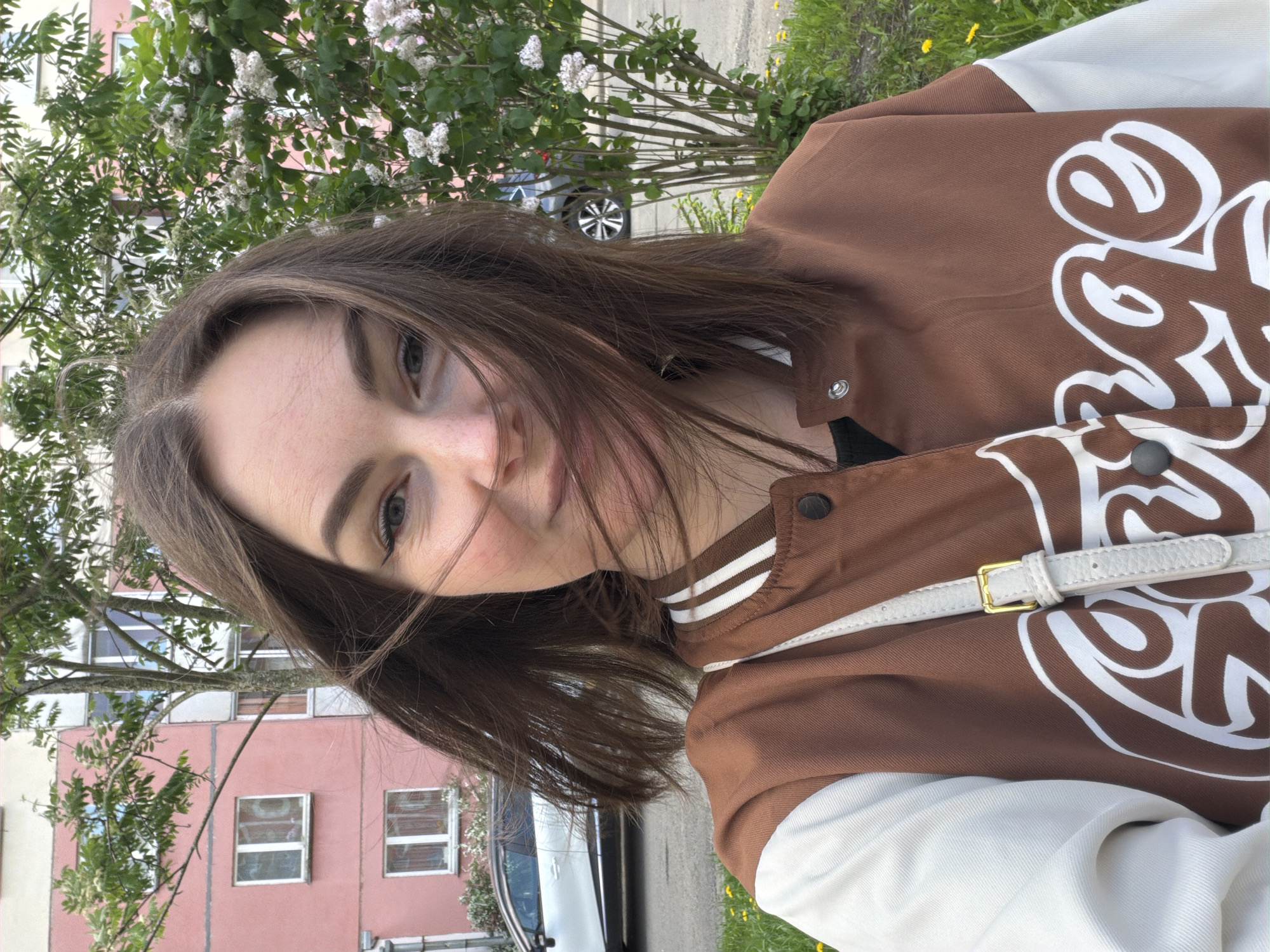 A person is angled in the image, showcasing their upper body in front of an urban landscape. They have medium-length, dark brown hair and are wearing a brown bomber jacket with white lettering on the right side and white striped details around the collar. A white shoulder strap is visible across their body. The person appears to be looking at the camera with a neutral expression.
In the background, there is a building with pink walls and white-framed windows on the left. A tree with white blossoms is overhead. A car is parked along the street, which is partially visible. Green grass is seen on the ground, suggesting a grassy area beside the road.
