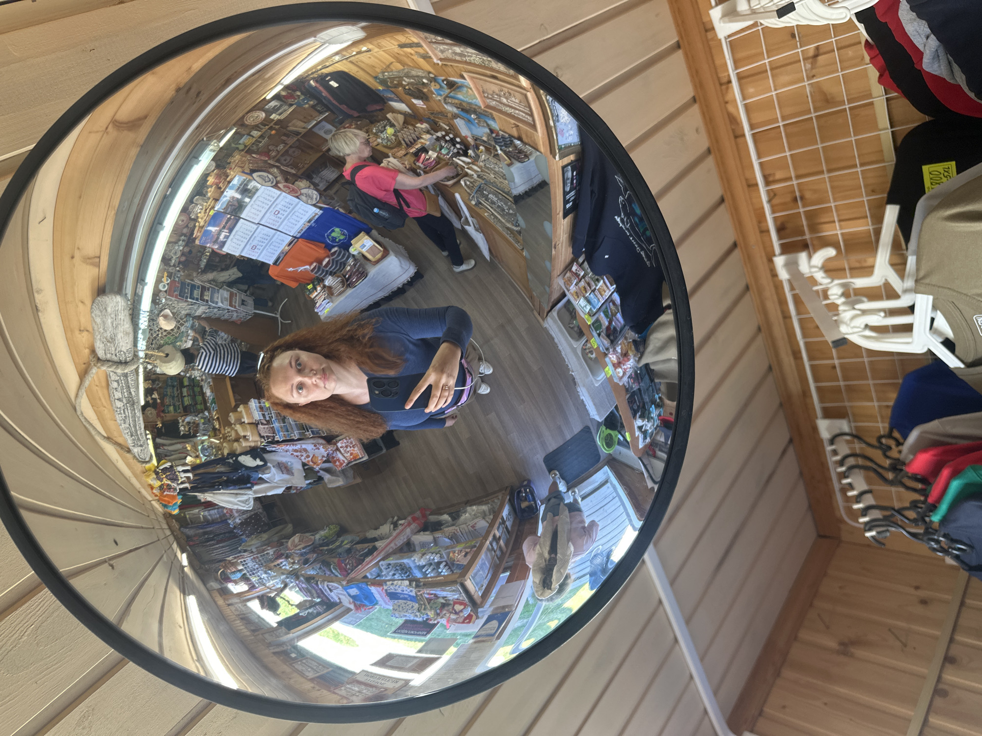 A distorted reflection in a convex mirror captures a woman with red hair taking a selfie. She is wearing a blue long-sleeved shirt and white sneakers, holding a smartphone in her right hand. The mirror reflects a small shop interior filled with shelves of merchandise, including textiles, souvenirs, and calendars. Another woman with blonde hair, wearing a pink shirt and backpack, is visible browsing the shelves in the reflection. The shop appears to be a souvenir store, possibly located in Solovetskiy, Russia, given the context provided. The area outside the mirror shows wooden walls and hanging clothes.