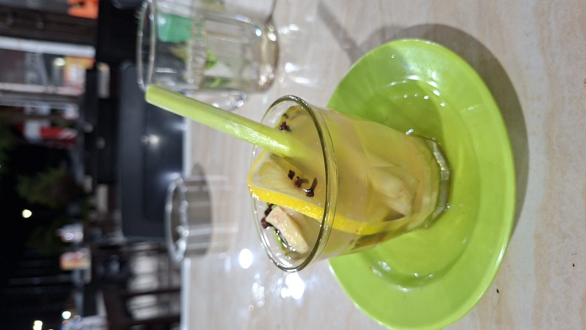 A close-up shot captures a glass of a yellow beverage on a bright green saucer. The drink contains slices of lemon, ginger, and a stalk of lemongrass, which extends out of the glass. The beverage appears to be a refreshing herbal drink. In the background, blurry out-of-focus elements suggest an outdoor seating area, possibly at a cafe or restaurant. The lighting indicates it is nighttime. The table surface is light-colored with a subtle patterned texture. An ashtray is visible in the background, suggesting a smoking-friendly environment. The image provides no specific clues to pinpoint the exact location within Keuniree, Indonesia, beyond it being an establishment with outdoor seating.