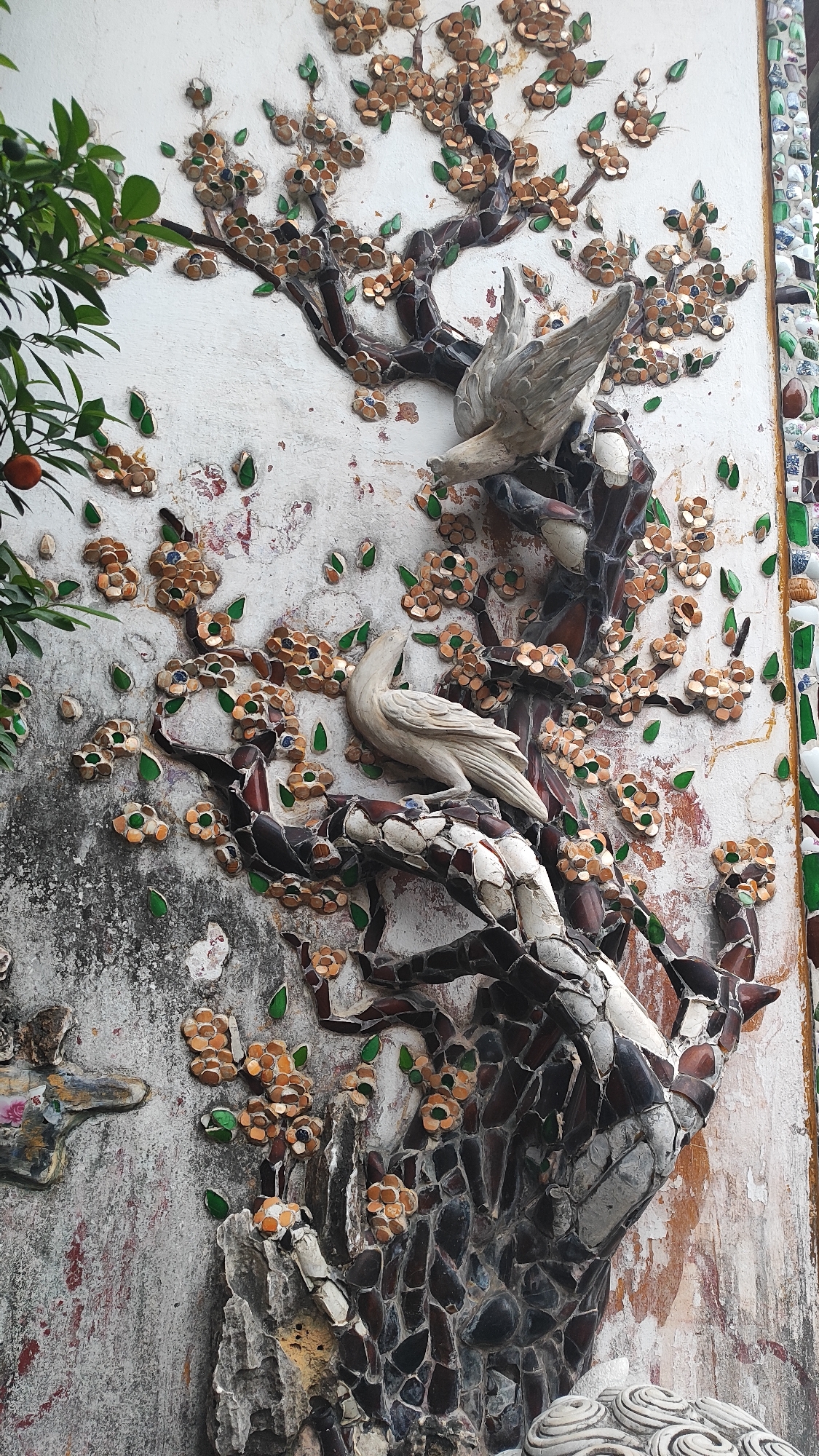 The image shows a detailed relief sculpture on a wall, depicting a stylized tree with birds and flowers. The tree's trunk and branches are made of dark, irregular pieces, possibly glass or ceramic shards, set against a textured, off-white wall. The branches are adorned with clusters of small, flower-like ornaments, each with a brown center and petal-like pieces, interspersed with small green leaf-shaped elements. Two white, bird-like figures are integrated into the sculpture. One bird, near the top, appears to be in flight or perched with its wings partially extended. The second bird, lower down, is perched on a branch, looking to its left with its head slightly tilted. The wall itself shows signs of age and weathering, with some patches of peeling paint and discoloration. To the left of the main sculpture, a real branch with green leaves and a single small orange fruit is visible, suggesting the sculpture is outdoors and near actual foliage. The overall style of the artwork, particularly the use of broken ceramic or glass pieces to form the relief, is characteristic of decorative art found in some Vietnamese temples and pagodas, such as those in the city of Đà Nẵng. Specifically, this style is commonly seen in Buddhist temples in Vietnam, often incorporating detailed mosaics and reliefs.