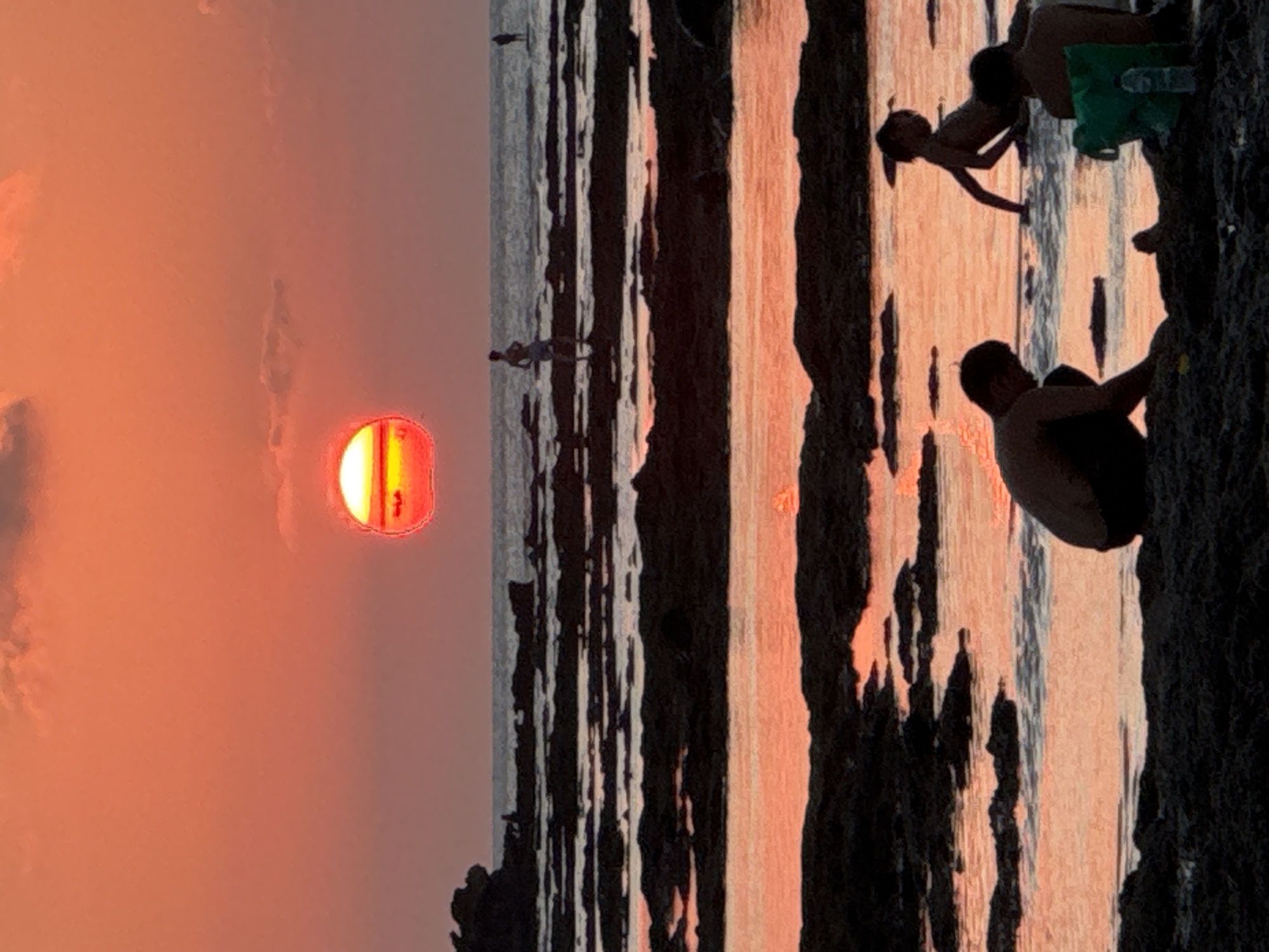 A group of people are at the beach in Kuta Selatan, Indonesia, as the sun sets over the ocean. The sky is a vibrant orange and pink, with scattered clouds. The sun, a fiery orb, is partially obscured by the horizon. In the foreground, several people are silhouetted against the bright sky. One person is kneeling and digging in the sand. Two children are playing in the shallow water, and another person is seated. In the distance, two more individuals are standing on the rocky shoreline, looking out at the sea. The water is calm and reflects the colors of the sunset. Rocks jut out of the water, creating a textured landscape. A green bag and a water bottle are visible on the sand in the foreground.