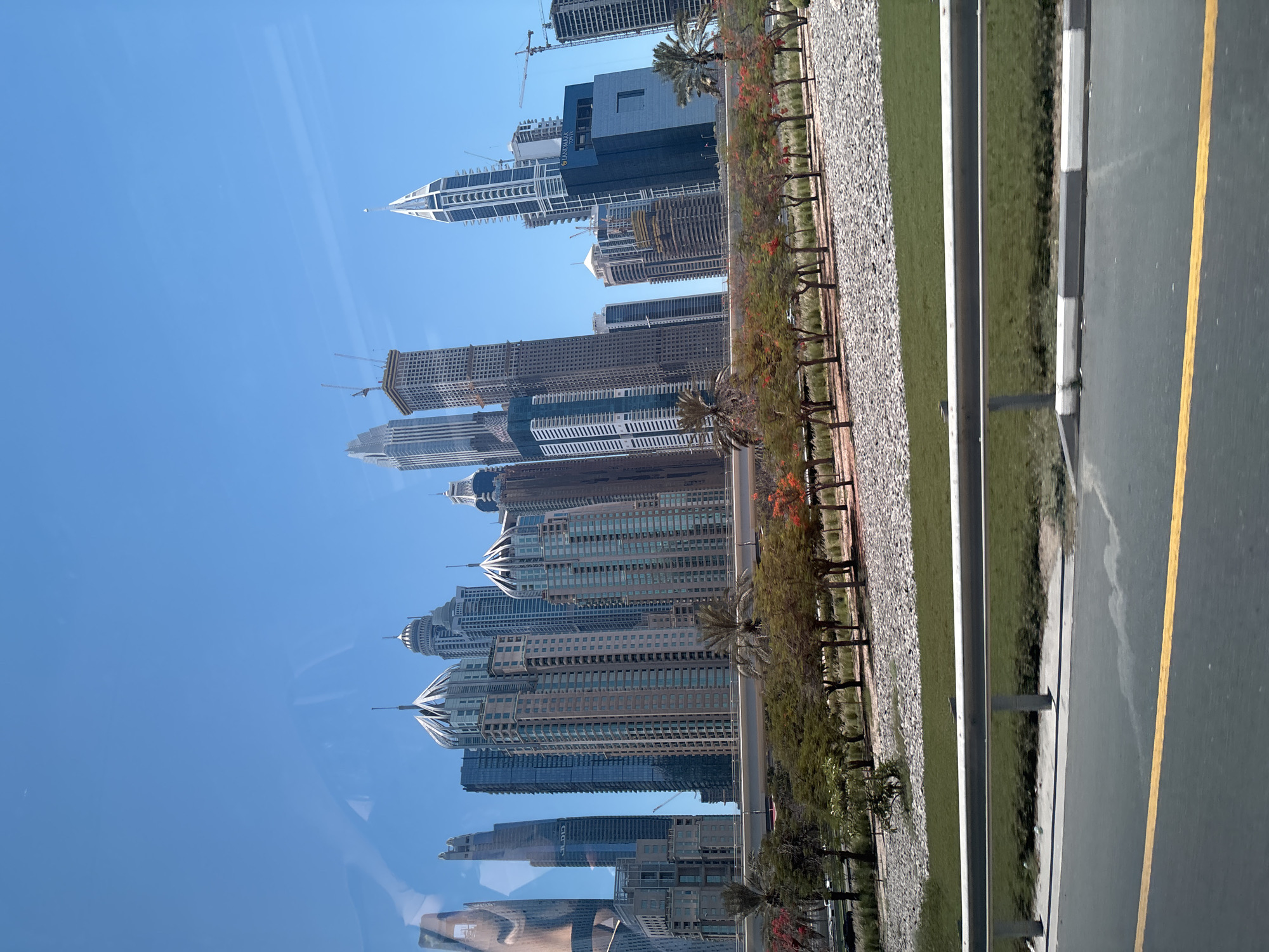 The image captures a view of Dubai Marina in Dubai, United Arab Emirates, showcasing a cluster of modern skyscrapers under a clear blue sky. In the foreground, a highway or elevated road runs horizontally across the frame, with a metallic guardrail separating it from a landscaped area. This landscaped area features a row of trees with sparse green foliage and some red flowers, interspersed with palm trees and a ground cover of grey pebbles and green grass. The iconic skyscrapers of Dubai Marina, with their varied architectural designs and heights, dominate the background. Some of the buildings appear to be under construction, indicated by cranes visible at their summits. The overall scene depicts an urban environment characterized by contemporary architecture and meticulously maintained green spaces, typical of Dubai's developed areas.