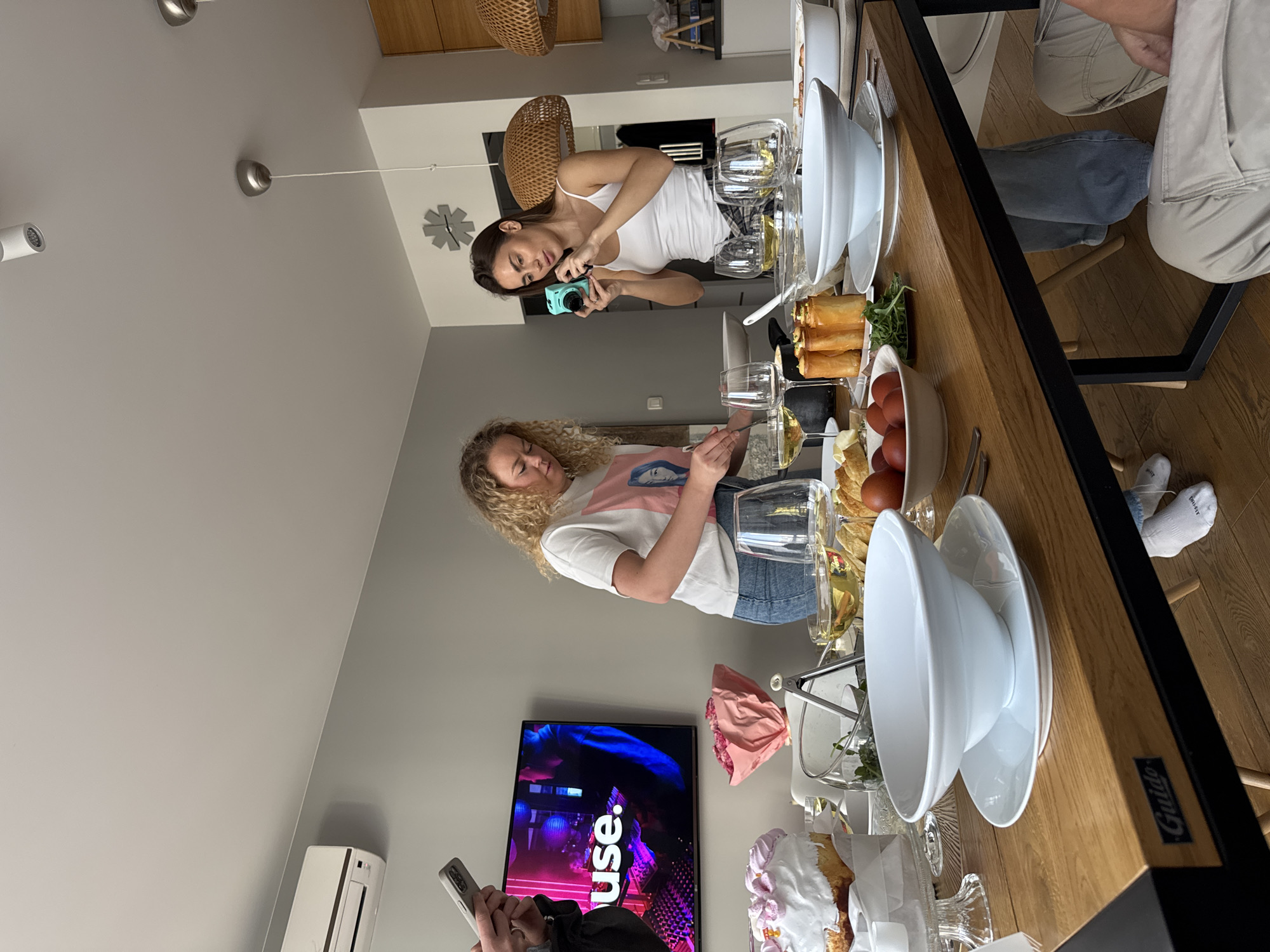 The image depicts an indoor scene with several individuals gathered around a table laden with food. Two women stand on opposite sides of the table. The woman on the right is using a turquoise camera to take a picture, while the other woman is holding a wine glass. On the table are various food items, including eggs, pastries, and salads, along with wine glasses and serving dishes. A television displaying the word "use" in large letters is visible in the background. The environment appears to be a well-lit apartment with modern decor. The presence of the text "Guido" on the edge of the table suggests the image was taken in the city of Warsaw, Poland, as it is a brand known to be sold in Poland.