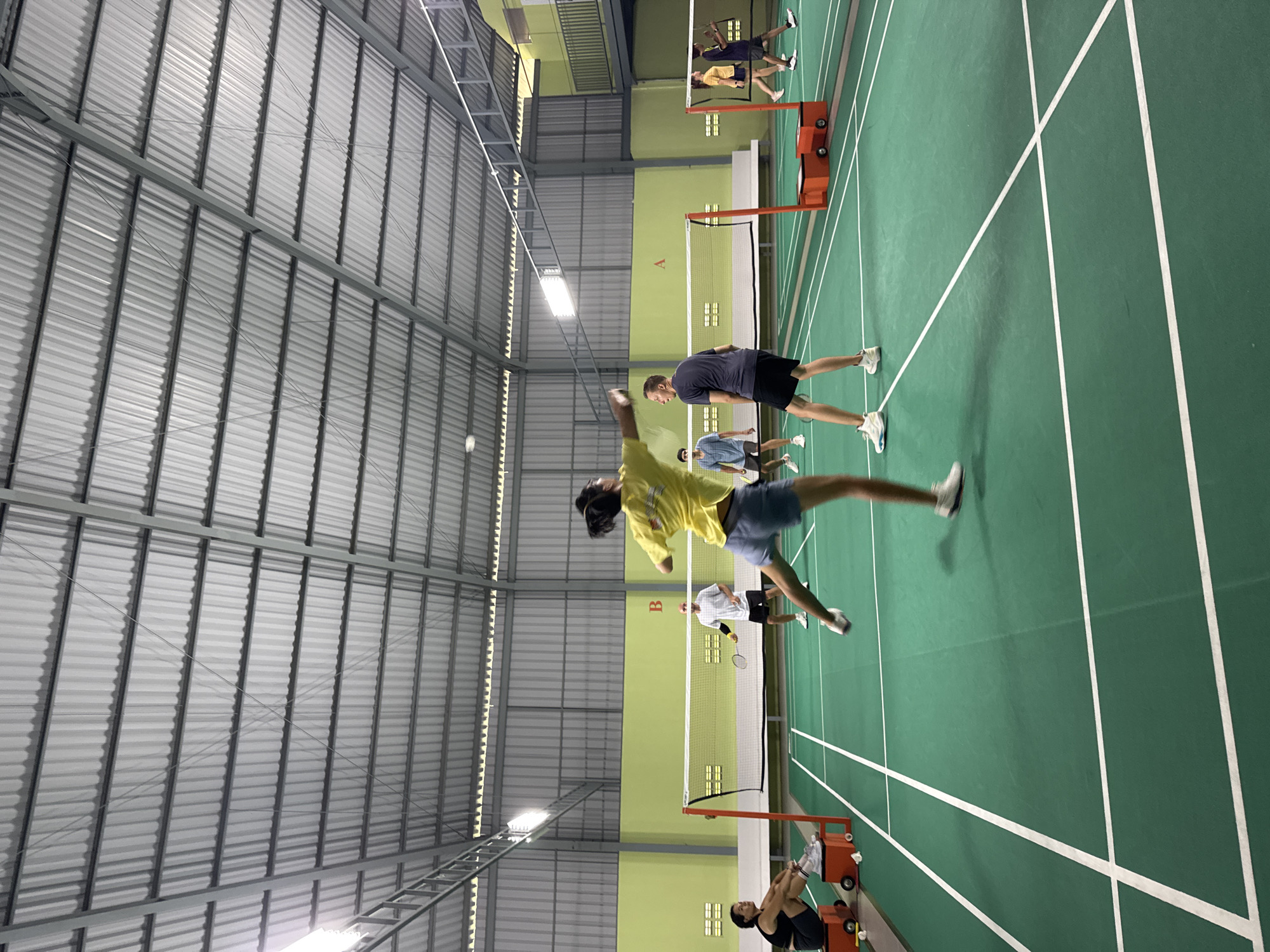 A badminton court, possibly indoors, is depicted with players engaged in the sport. One player wearing a yellow shirt is leaping to hit the shuttlecock. There are multiple other players distributed across the green court, defined by white lines and separated by a net. The setting appears to be a large, industrial-style indoor space with a high ceiling made of corrugated metal, illuminated by overhead lights. Specific location details within Unknown, Unknown, are not discernible from the image.