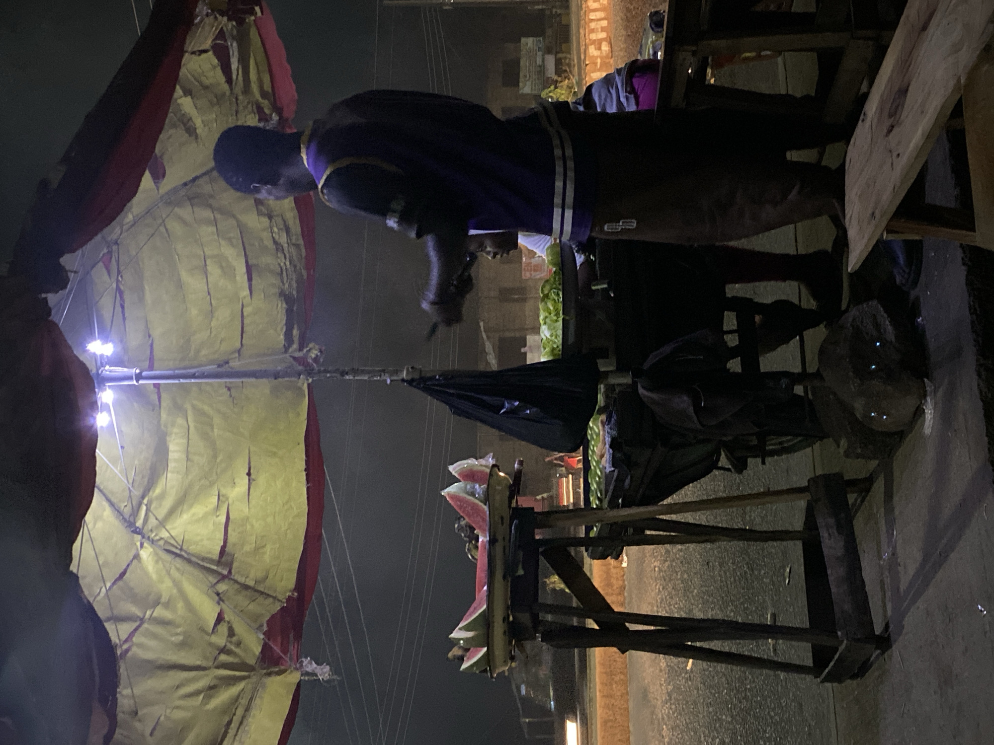 This image depicts a nighttime street market scene, likely in Bukuru, Nigeria, featuring a vendor, their produce stall, and a prominent canopy.
In the center, a vendor, dressed in a dark purple top with light-colored stripes, stands behind a wooden stall. They appear to be engaged in commercial activity, possibly serving a customer or arranging their goods. The stall displays various fresh items, most notably several large, vibrant pink slices of watermelon arranged on its top surface. Below these, bundles of green leafy vegetables, resembling lettuce or cabbage, are also visible.
Above the vendor and stall, a large canopy or umbrella provides shelter. Its fabric, a combination of faded yellow and red, appears worn and tattered in several places. Multiple small, bright artificial lights are attached to the canopy's frame, casting a strong, localized illumination on the produce and the immediate area of the stall, contrasting sharply with the general darkness of the night.
The background is largely dark and indistinct, with faint outlines of structures suggesting other market elements or buildings under the night sky. The ground beneath the stall appears to be a paved surface. The scene conveys the atmosphere of active nocturnal commerce. No legible text is discernible within the image.