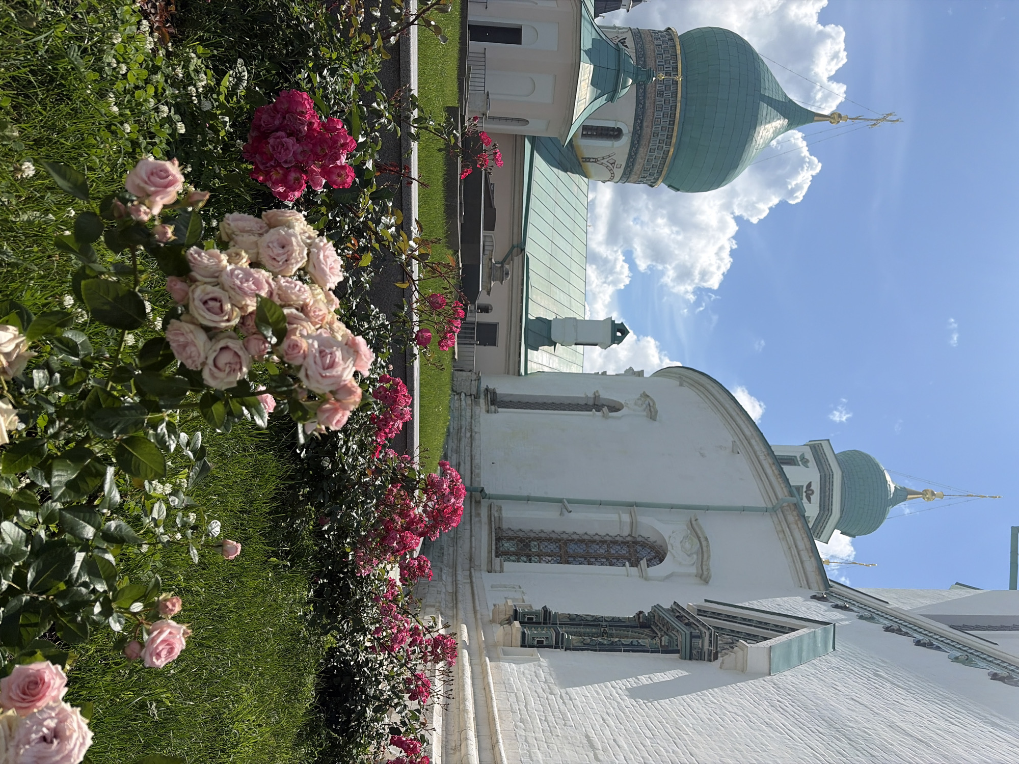 This is a medium shot, eye-level photograph of a garden with blooming roses in the foreground and a historic white building with green domes in the background. The building appears to be a religious structure, possibly a monastery or church, with ornate windows and a gilded cross atop one of the domes. The roses are in various shades of pink and red, with lush green foliage. The sky is blue with fluffy white clouds. The scene is well-lit, suggesting it was taken on a sunny day. The location is the Voskresensky New Jerusalem Monastery in Istra, Russia. There are no people or animals visible in the image.