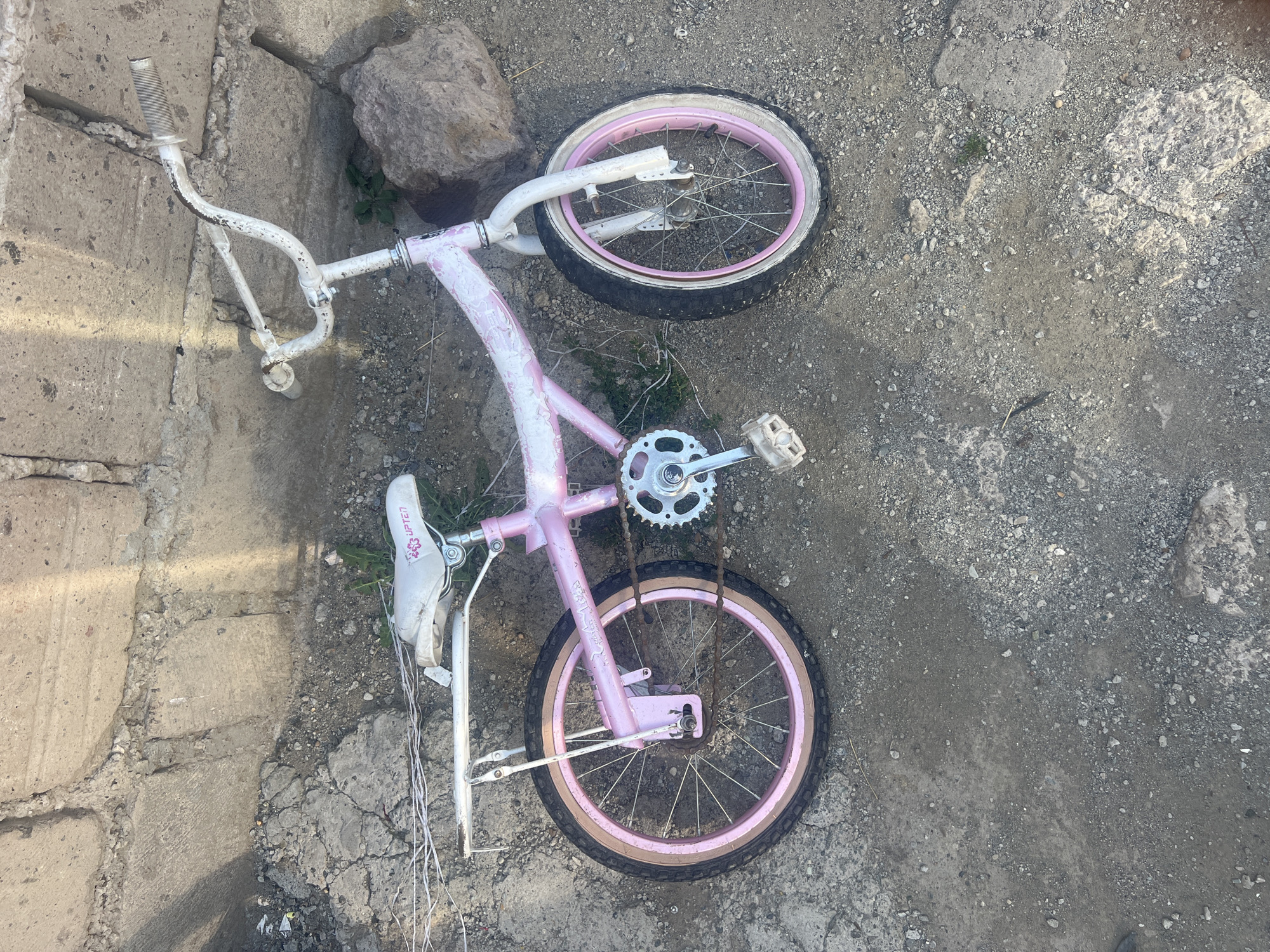 A pink and white children's bicycle lies on its side on a rough, sandy ground next to a concrete wall. The bicycle has a white seat with a pink logo, white handlebars, and pink rims on both wheels. The bicycle appears to be stationary and abandoned. The concrete wall is made of large, rough-hewn stones. There is some dry vegetation and debris scattered around the scene.