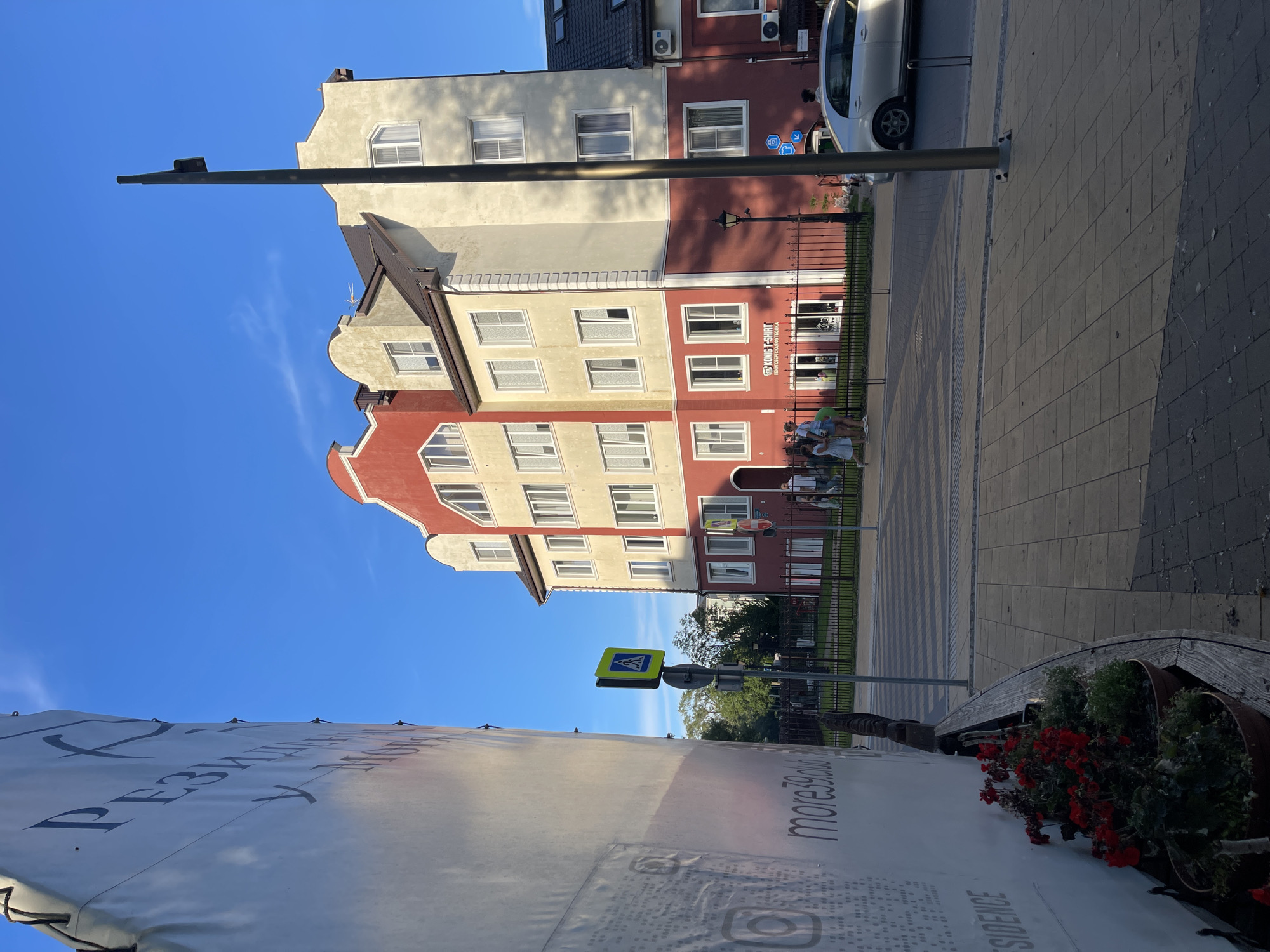 The image depicts a street scene in Zelenogradsk, Russia, near the intersection of Sovetskaya Street and Kurortny Prospekt. On the left, a large white banner with dark lettering is partially visible, with the text "more39.club" and "RESIDENCE" discernible. In the center, a multi-story building features a distinctive red and cream facade with white-framed windows. A sign reading "KONIG T-SHIRT" is visible on the ground floor. A pedestrian crossing is marked on the paved sidewalk, with a yellow pedestrian crossing sign in place. A thin lamppost stands to the right of the building. A silver car is parked on the street. Several individuals are seen walking on the sidewalk and near the building entrance. Red flowers are planted in pots along a curved wooden bench in the foreground. The sky is clear blue with a few wispy clouds.