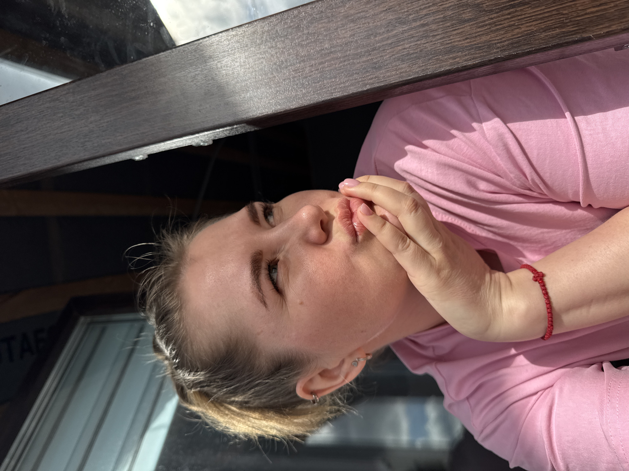 A woman with light skin and blonde hair is shown from a low-angle perspective. She is wearing a pink long-sleeved shirt and has a red string bracelet on her left wrist. Her hands are near her mouth, and she appears to be resting her face on them. She has multiple piercings in her left ear. The background includes a dark wood-like structure and a window with writing. The location is Gornyi (Dvurechenskoe S/P), Russia.
