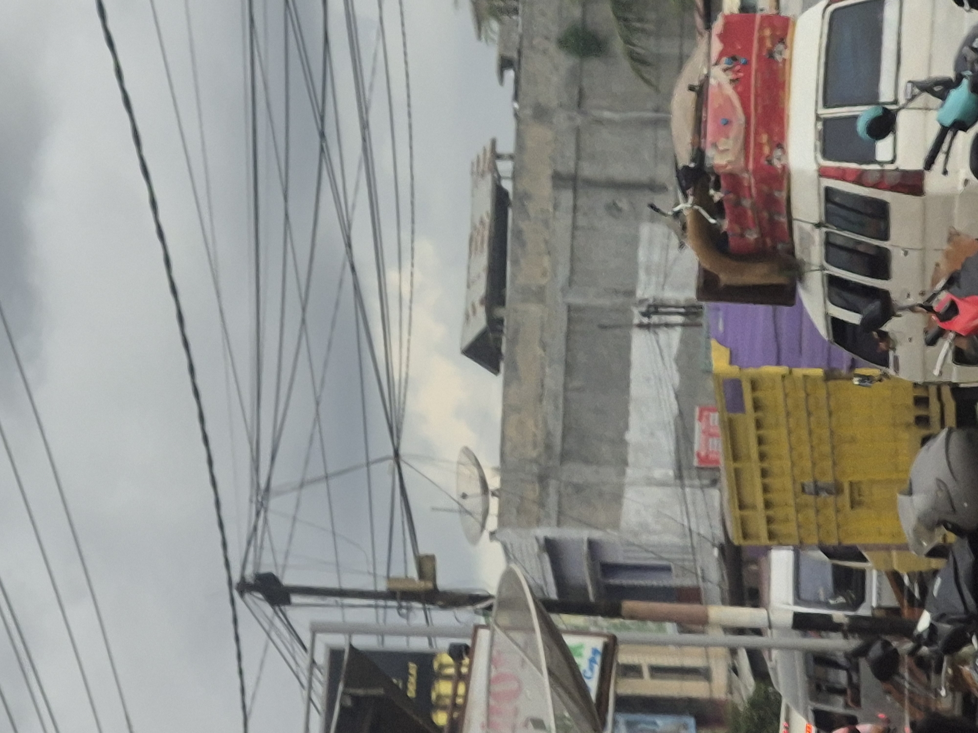 The image depicts a street scene in Tingkeummanyang, Indonesia. Overhead power lines crisscross the sky. Buildings and commercial establishments line the street, including structures with satellite dishes. Vehicles, including motorcycles and auto rickshaws, are present. A person wearing a helmet is visible on a motorcycle.