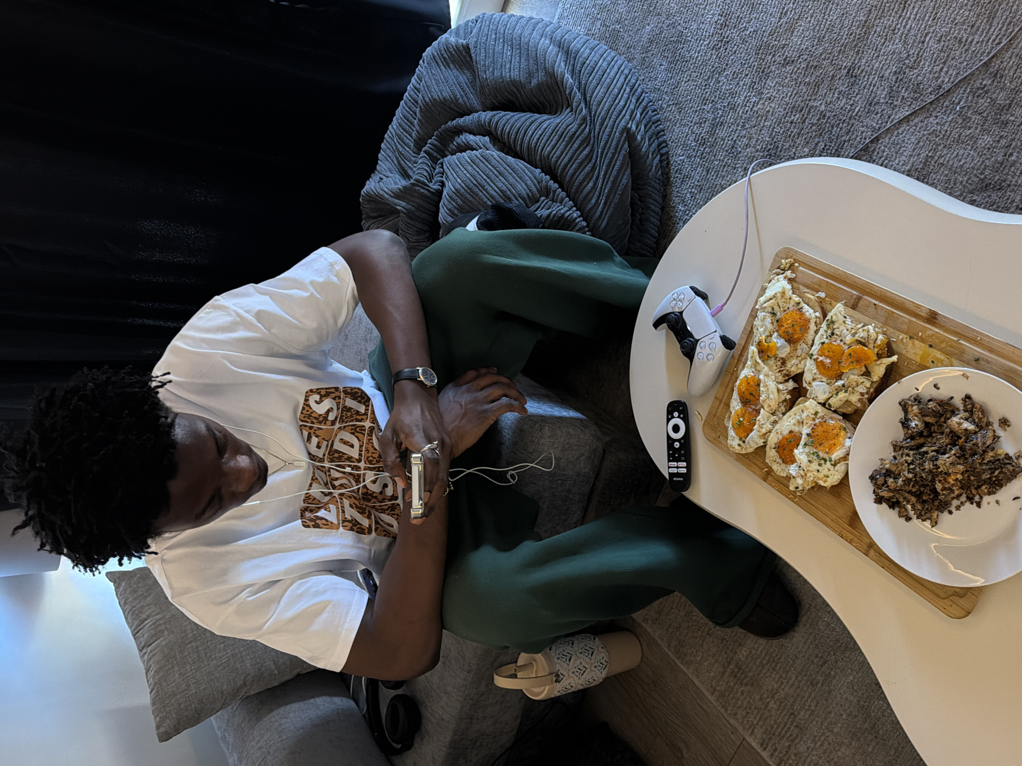 A young Black man with dark, curly hair is seated on a grey couch, looking down at his smartphone, which he holds in his left hand. He is wearing a white t-shirt with a graphic print, green sweatpants, and black slip-on shoes. White wired earphones are connected to his phone and draped around his neck. He wears a silver ring on his left index finger and a watch on his left wrist.
In front of him is a white, oval-shaped coffee table. On the table, a wooden cutting board holds four pieces of toast topped with fried eggs and chives. To the left of the toast is a white plate with a pile of dark, crumbled food, possibly mushrooms or meat. A white PlayStation controller, connected to a purple cable, rests on the table next to the toast. A black remote control with circular buttons is also on the table.
Behind the man, a large, grey, corduroy beanbag chair is visible. To the left of the couch, a grey cushion is placed against the back of the sofa. Near the floor, on the left side of the frame, is a beige cylindrical object with a woven pattern. The floor is covered with a grey rug, and a light-colored wooden floor is visible in the background. The overall scene suggests a casual indoor setting. There is no indication of the location being in the city of London, United Kingdom.
