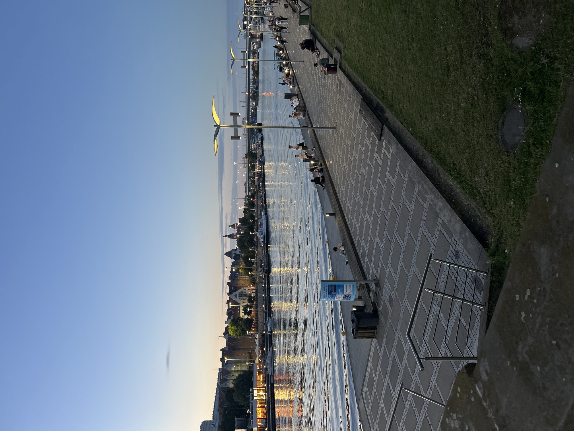 This image captures an evening scene along the Oder River waterfront in Szczecin, Poland. The water reflects the warm glow of lights from buildings on the opposite bank, which feature a mix of traditional architecture with red roofs and larger, modern structures. Several boats are docked or moving on the river.
The foreground shows a wide paved promenade with a distinctive geometric pattern of light and dark grey tiles. Several lampposts with distinctive curved, illuminated shades line the promenade, casting a warm light. A grassy area with a few scattered flowers runs alongside the promenade.
People are seen relaxing and strolling along the waterfront. Some are seated on benches or the edge of the promenade, while others walk further down. There are trash receptacles and a blue information kiosk visible on the promenade.
In the distance, a bridge spans the river, and the city skyline is illuminated against a twilight sky that transitions from pale blue to a warmer hue near the horizon. The overall atmosphere is peaceful and serene, suggesting a popular gathering spot for locals and visitors.