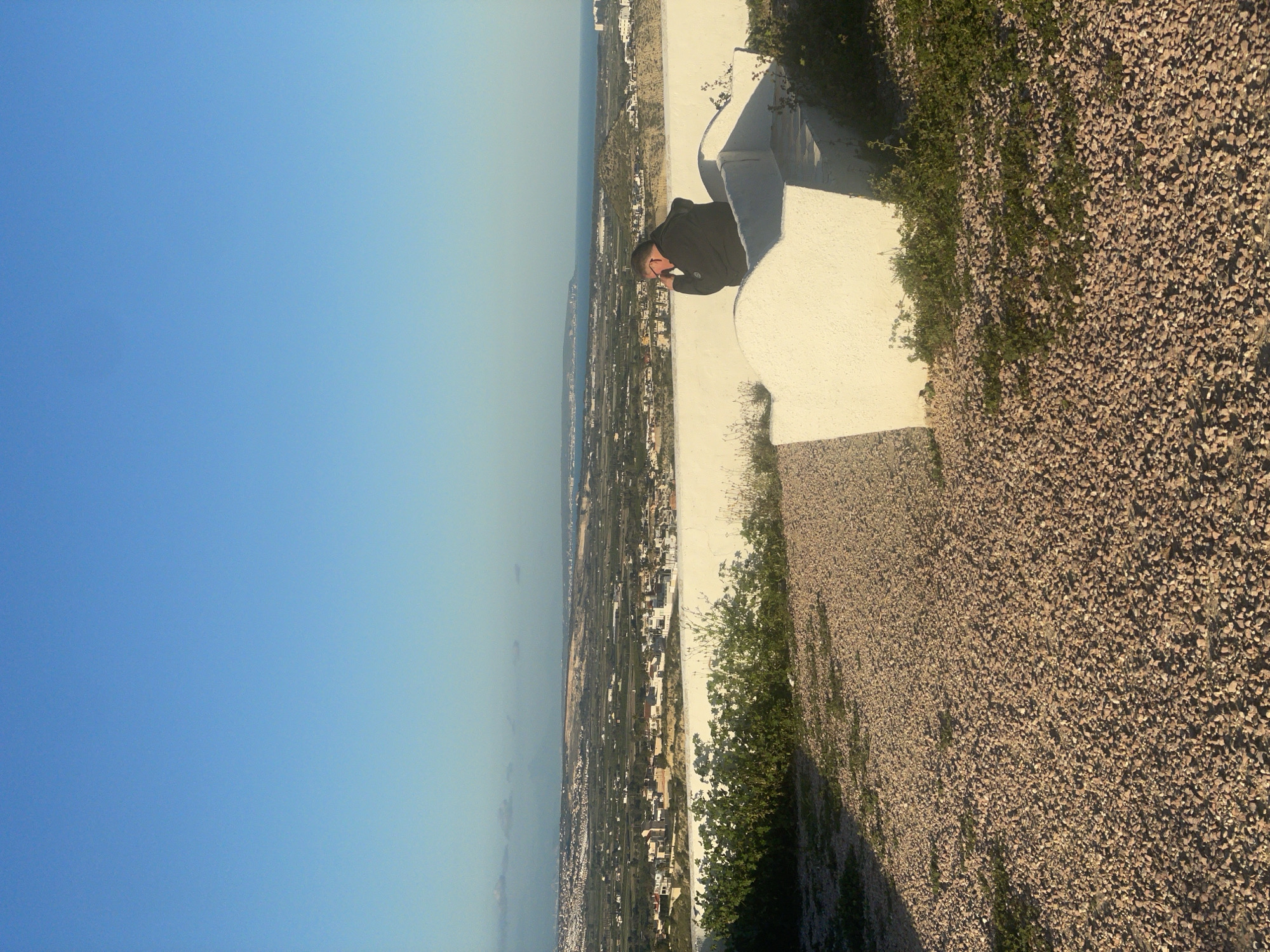 A man in a dark jacket leans against a low, white wall at a high vantage point overlooking a coastal urban area, specifically Rojales, Spain. The sky is blue, with a slight gradient. The ground to the right of the wall is covered in small, light-brown rocks interspersed with patches of greenery. The urban sprawl is visible between the wall and the sea.
