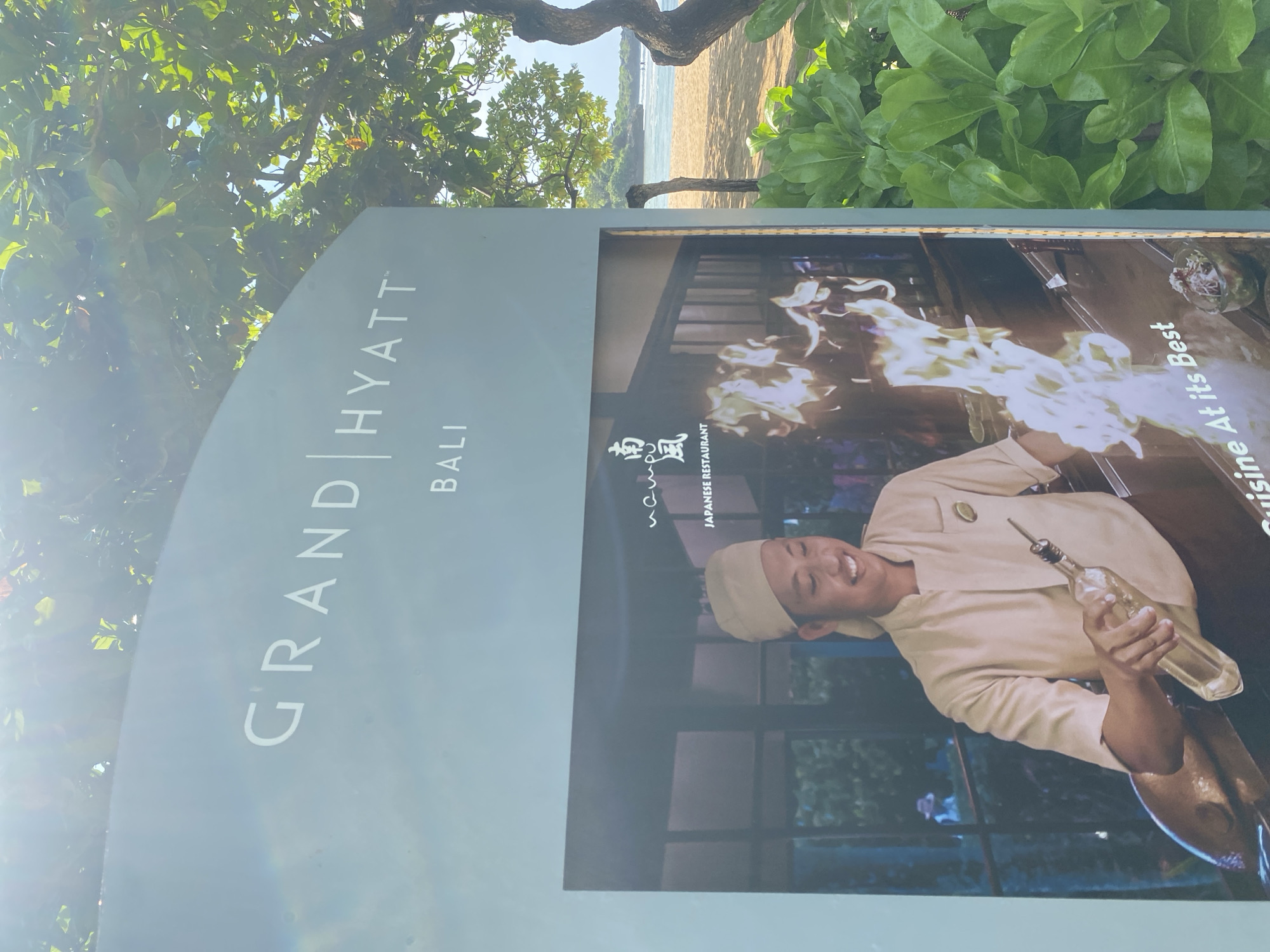 The image shows a sign for the Grand Hyatt Bali in Nusa Dua, Indonesia. The sign features the hotel logo and an advertisement for its Japanese restaurant, showcasing a smiling chef with a bottle in hand, set against a backdrop of flames. The photograph is taken outdoors, with trees and vegetation visible in the background. In the far background, the beach with sand and the ocean is visible. The image is taken during daytime with bright sunshine visible in the image.