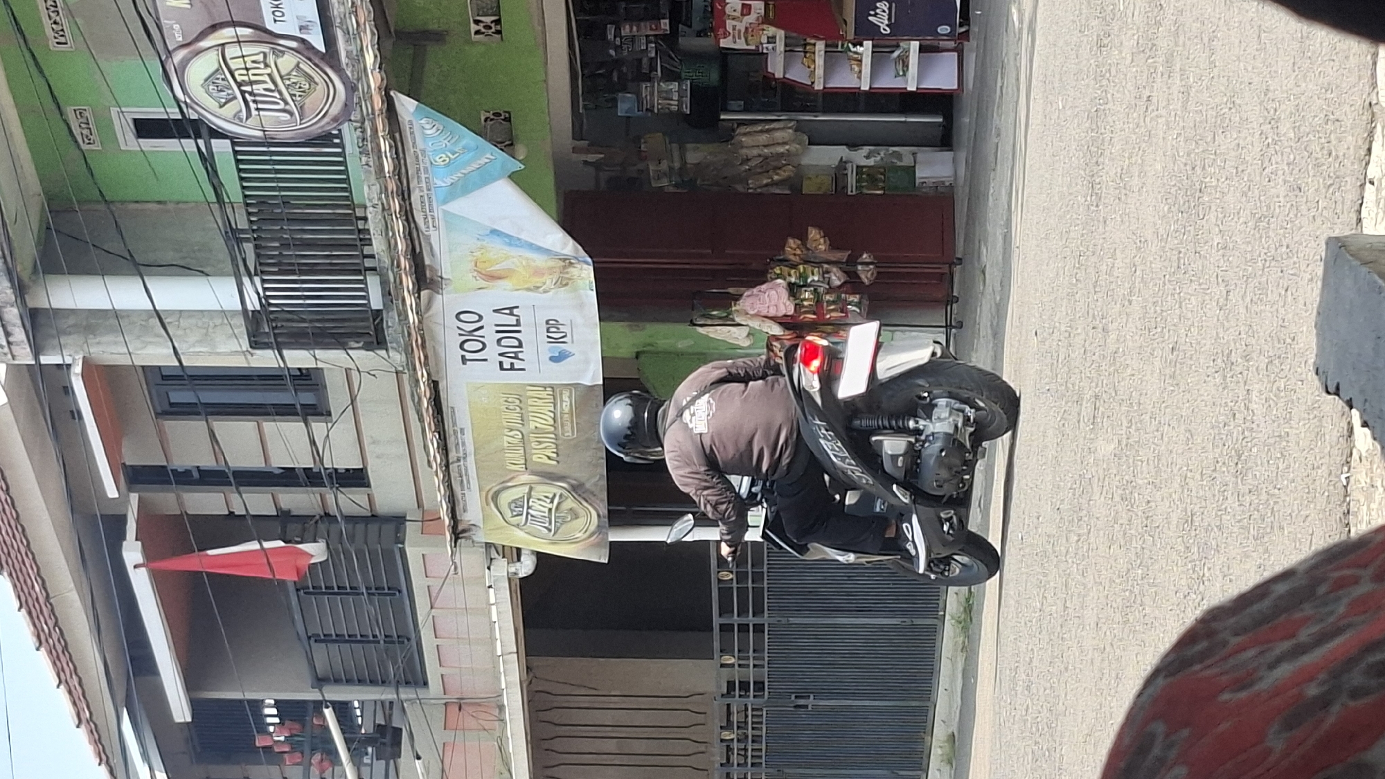 The image captures a street scene in Cilaku, Indonesia, likely a residential and commercial area. In the foreground, a person is riding a motorcycle, wearing a brown long-sleeved shirt and black pants, with a black helmet. The motorcycle is black and white. In the background, there are buildings with balconies, some adorned with the Indonesian flag. A banner with text including "TOKO FADILA" and "KPP" is displayed. Various shops are visible, one with a green facade and a circular logo featuring the word "JUARA". Another shop displays shelves with products, including items from "Alice". The street is paved and appears to be a sunny day.