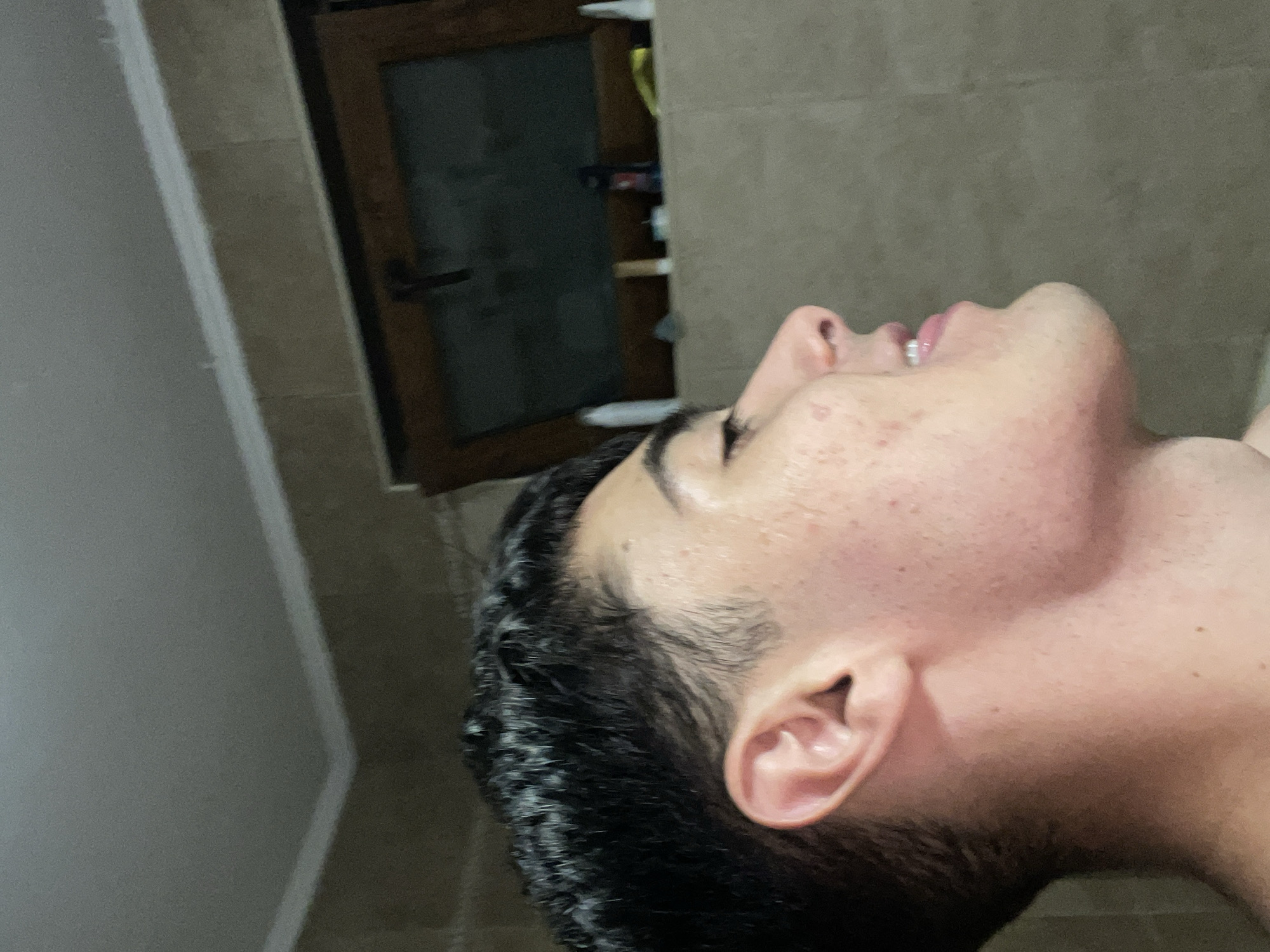 The primary subject of the image is a person, likely a young male, seen in a close-up profile view, facing upwards and towards the right of the frame. His head is tilted back, with his mouth slightly open, revealing teeth. He has dark, curly hair, prominent eyebrows, and visible skin texture, including some blemishes on his cheek and forehead.
The background suggests an indoor setting. To the left and behind the person's head, there's a light-colored, plain wall. To the right and further back, there are light brown or beige tiles on a wall, and a wooden door with a dark handle is partially visible. Behind the door, a faint outline of what appears to be shelves or a cabinet with objects on them can be discerned in some of the cropped hints.
The activity depicted is simply the person looking upwards with a neutral expression. No specific event is taking place. The lighting is artificial and somewhat dim, consistent with an indoor environment. There is no visible text in the image.