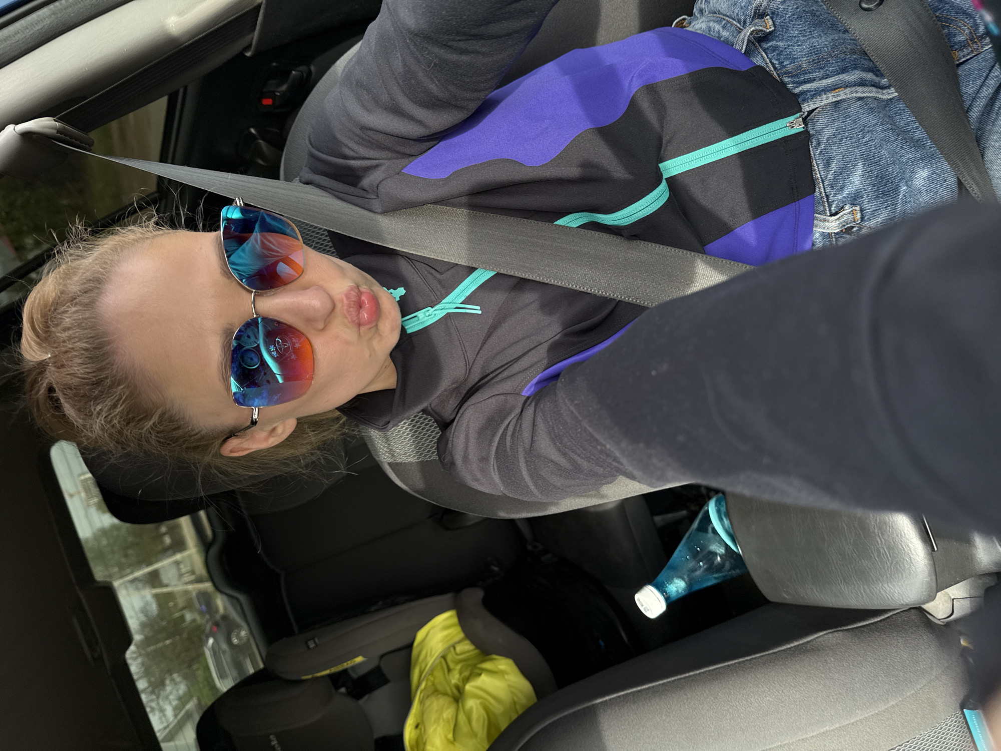 A woman with light hair pulled back into a ponytail, wearing blue and red reflective sunglasses, a black jacket with purple and turquoise accents, and blue jeans, is seated in a car and wearing a seatbelt. She is making a "kissing" expression. A water bottle and a yellow jacket are visible in the back seat. The location is Chaikovsky, Russia, as implied by the prompt, although this is not discernible from the image itself.