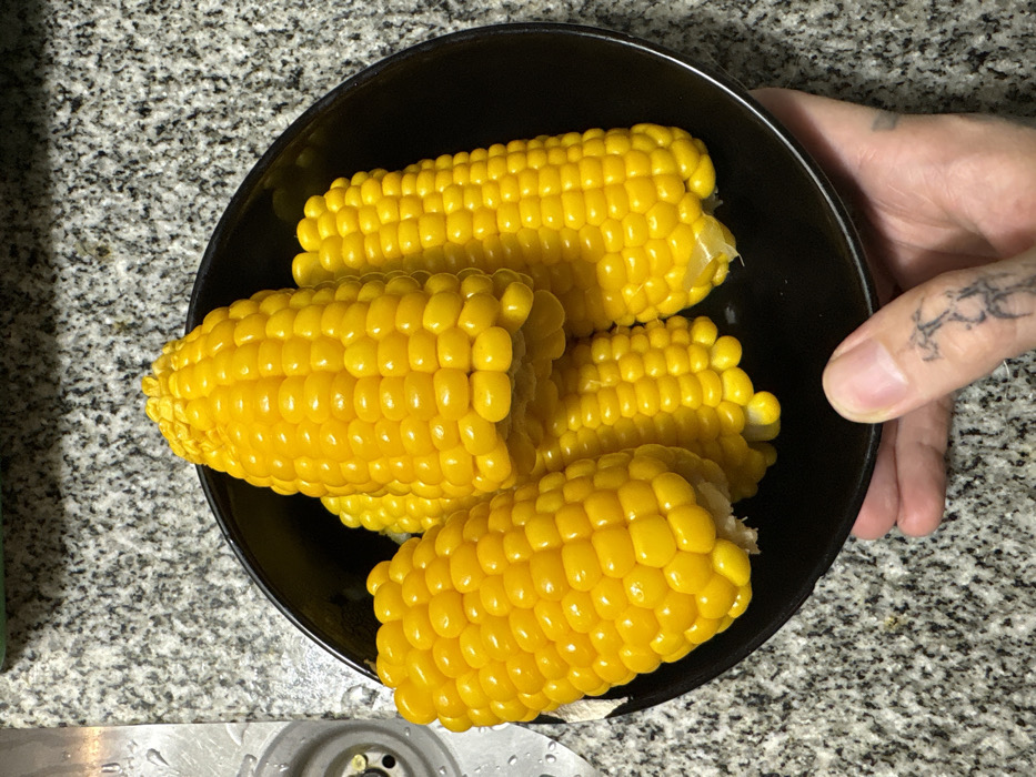 The scene depicts a close-up view within a domestic kitchen setting, specifically a countertop area. A black, circular bowl contains five cooked ears of yellow corn, visible with uniform, plump kernels. The corn is bright yellow, suggesting a fresh or recently prepared state. A human left hand, visible from the wrist down, supports the bowl from underneath and slightly to the right. The hand exhibits multiple tattoos: a prominent skeletal or abstract design on the dorsal side of the thumb and a smaller, similar design on the index finger. The fingernails are clean and trimmed. The background consists of a light grey speckled granite or similar stone countertop. To the immediate left, a portion of a stainless steel sink basin is visible, including a central drain opening and several water droplets on its surface. A faint green object is partially discernable at the top edge of the frame. The overall environment is consistent with a residential kitchen in Buenos Aires, Argentina, as per the contextual information provided, although no specific geographical identifiers are visually present in the image itself.
