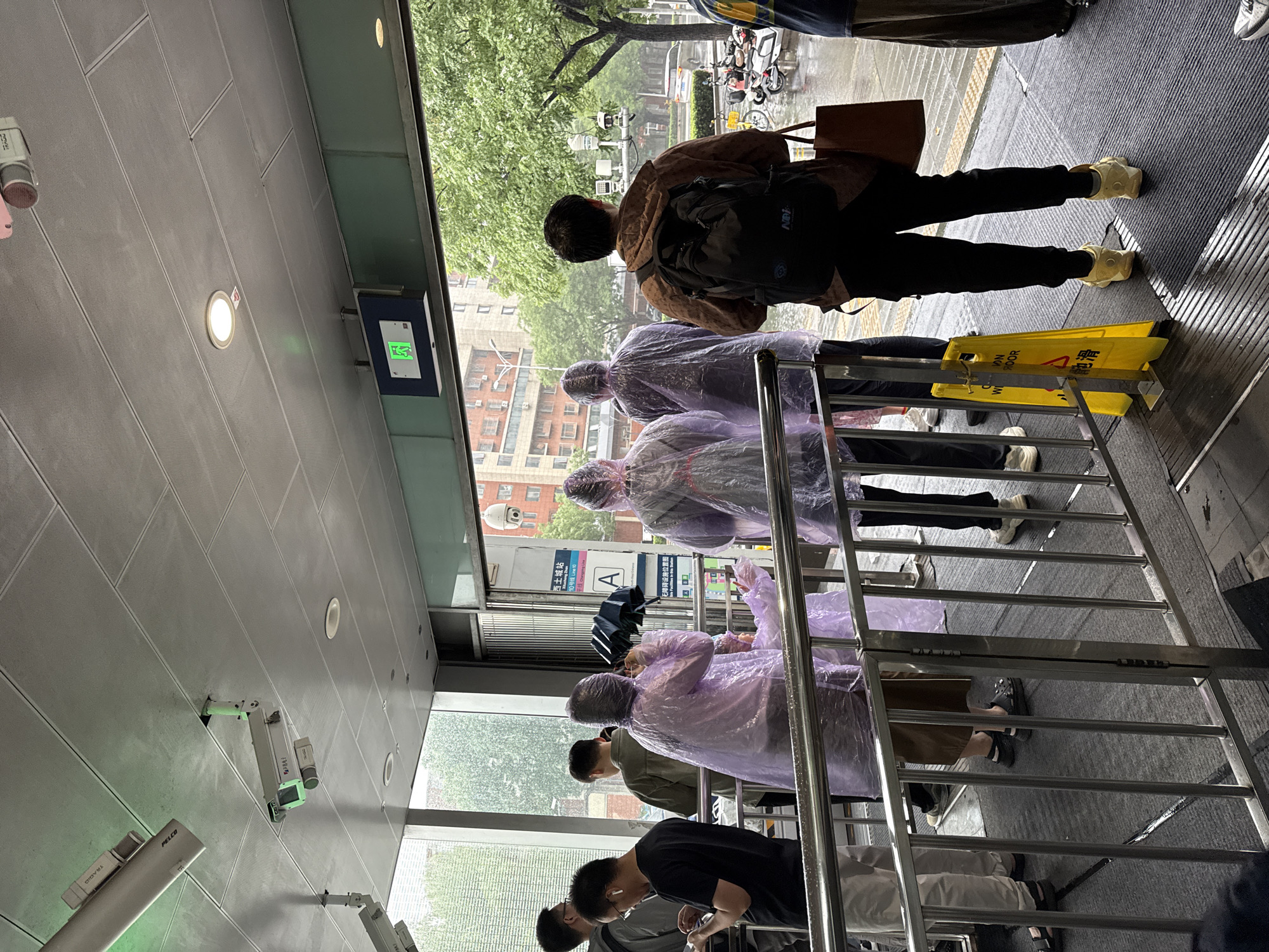 The image captures an indoor-outdoor transitional area at Exit A of Xitucheng Station, Line 10, Beijing Subway, Haidian District, Beijing, China, during rainy daylight conditions. The floor inside is covered with a dark grey, textured mat that appears wet in several places. A yellow A-frame "CAUTION WET FLOOR" sign with Chinese characters "小心地滑" stands to the right of a silver metal railing system that guides movement. The ceiling is composed of light grey rectangular panels, from which multiple white surveillance cameras (including brands "PELCO" and "TRACO") and recessed circular lights are mounted. A rectangular illuminated green exit sign, featuring a walking person symbol, is positioned above the entryway.
Approximately eight individuals are visible. Several people are wearing translucent light purple rain ponchos. One person, positioned centrally with their back to the viewer, wears a brown patterned jacket, black pants, and a black backpack, holding a brown tote bag. This individual stands on the wet floor mat near the yellow sign. To their left, two people in purple ponchos are seen from behind; one holds a closed dark umbrella. A smaller person in a purple poncho is beside them. To the far left, a person wearing a black t-shirt, glasses, and white earbuds is looking at a mobile phone. Another individual to their right wears a dark olive-colored jacket, facing away. A person in a blue and yellow striped shirt is partially visible on the far right.
The exterior view through large glass windows reveals wet asphalt, a vibrant green tree, residential buildings, parked motorcycles and scooters, and a pole-mounted weather monitoring station. The overall scene suggests people are either entering the station to escape the rain or exiting into it.