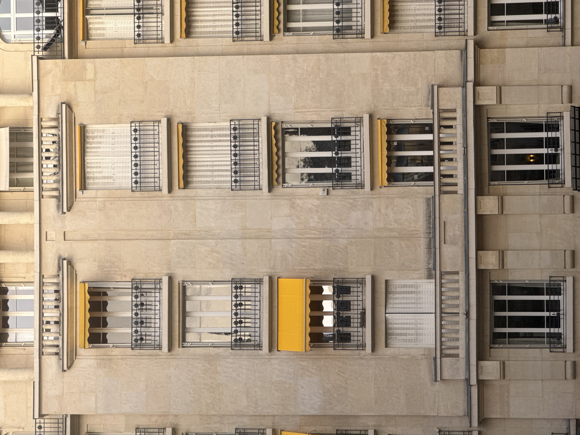 The image depicts the facade of a building in Paris, France. The building has a light beige or tan color and features a series of rectangular windows arranged in a grid-like pattern. Each window is adorned with a black metal railing or grill on the outside. Some windows have either white, striped shades, or yellow canopies partially drawn down, while others are open, revealing the interior. The architectural style of the building appears to be traditional Parisian, with distinct lines and features characteristic of the city's architecture. The overall impression is one of classic urban elegance.
