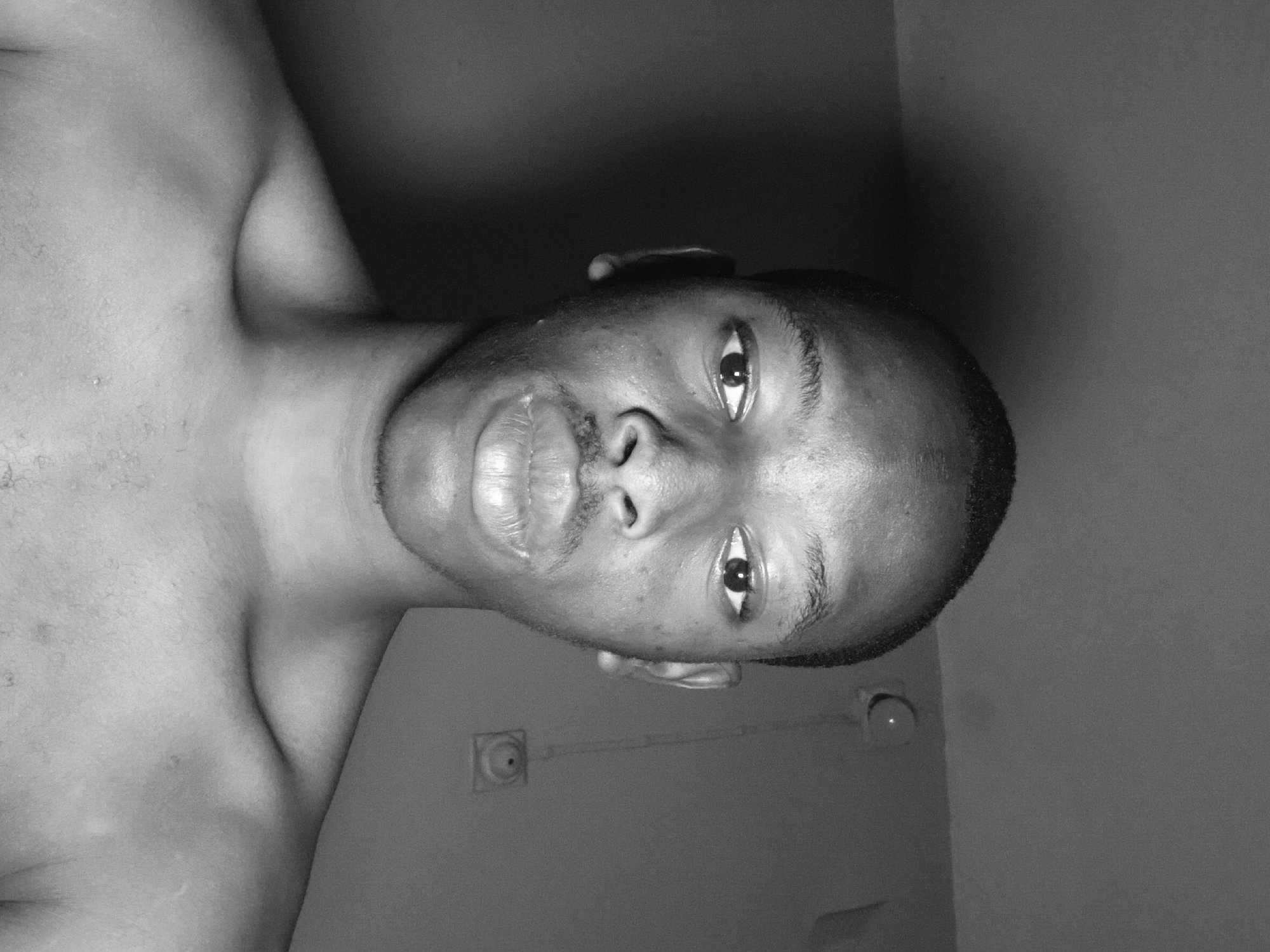 The image is a black and white, close-up, frontal portrait of a young Black man. He is looking directly at the camera with a neutral expression. His head is shaved, and he has a mustache. His shoulders are bare. The background appears to be a plain wall with some light fixtures. The image is likely taken indoors in an urban environment, possibly in Lagos, Nigeria.