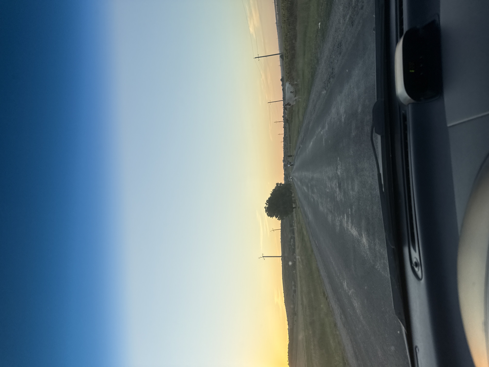 The image depicts a view from inside a vehicle, looking out at a rural road at dusk. The sky transitions from a deep blue at the top to a soft yellow and orange near the horizon, indicating sunset or sunrise. The road is paved but appears somewhat rough, with patches of lighter material. On either side of the road are grassy fields and vegetation.
In the distance, there are utility poles with wires stretching across the landscape. Several trees are visible, including a larger, fuller tree on the right side of the road. There are also a few indistinct figures walking on the side of the road and some animals, possibly cows, grazing or standing in the fields. A white vehicle is parked on the left side of the road in the distance. The foreground shows the dashboard of the vehicle, including part of the steering wheel and a small electronic device with a green light.
The scene is characteristic of a rural area, and the presence of utility poles and the type of vegetation suggest a setting in Moldova, specifically within or near the village of Cioc-Maidan. The overall atmosphere is calm and tranquil, with the fading light creating a sense of quietness.