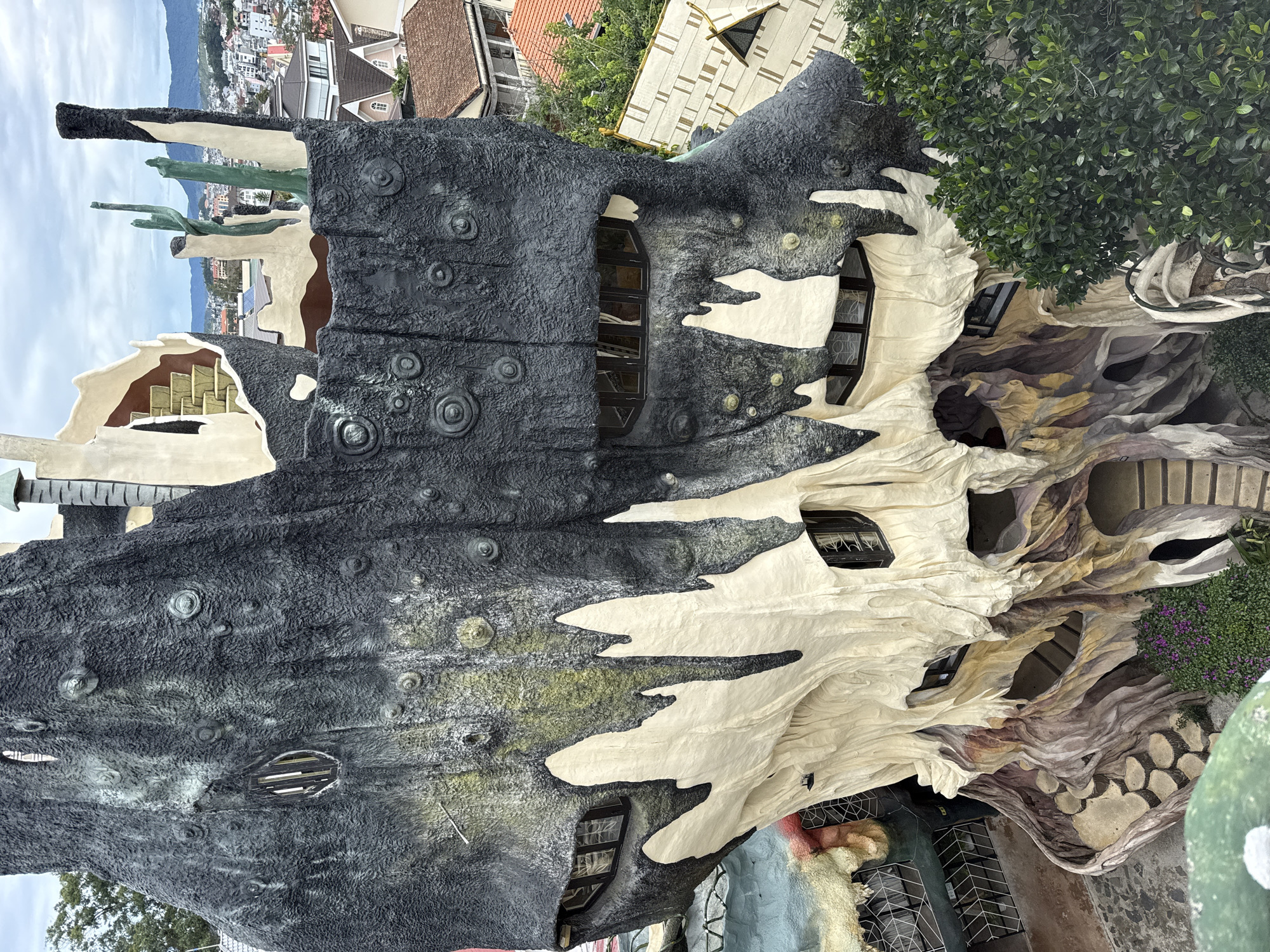The image shows Hang Nga guesthouse, also known as the Crazy House, in Da Lat, Vietnam. The building's exterior has an organic and surreal design, resembling a large tree or cave. The building is constructed with textured grey walls with embedded circular elements. There are white areas with irregular edges that extend around the structure. Windows and other openings are incorporated into the design. Nearby buildings are visible in the background.