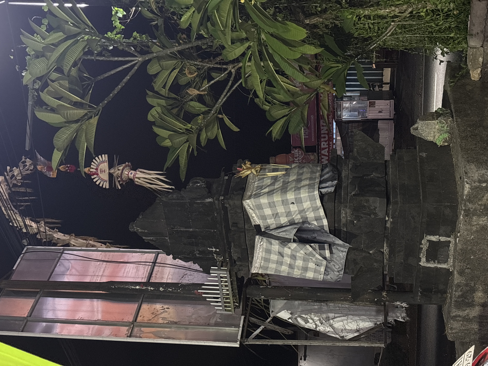 In Kuta Selatan, Indonesia, at night, a Balinese Hindu shrine is visible. The shrine is made of dark stone and is adorned with checkered cloth. Above it, traditional Balinese decorations hang. A lush green tree with large leaves stands to the right of the shrine. In the background, a brightly lit building with a sign that reads "WARUNG" is visible, suggesting a local food stall or small restaurant. The street is visible in front of the shrine, with some light reflecting off its surface. The overall scene depicts a typical roadside shrine in a commercial area of Bali.