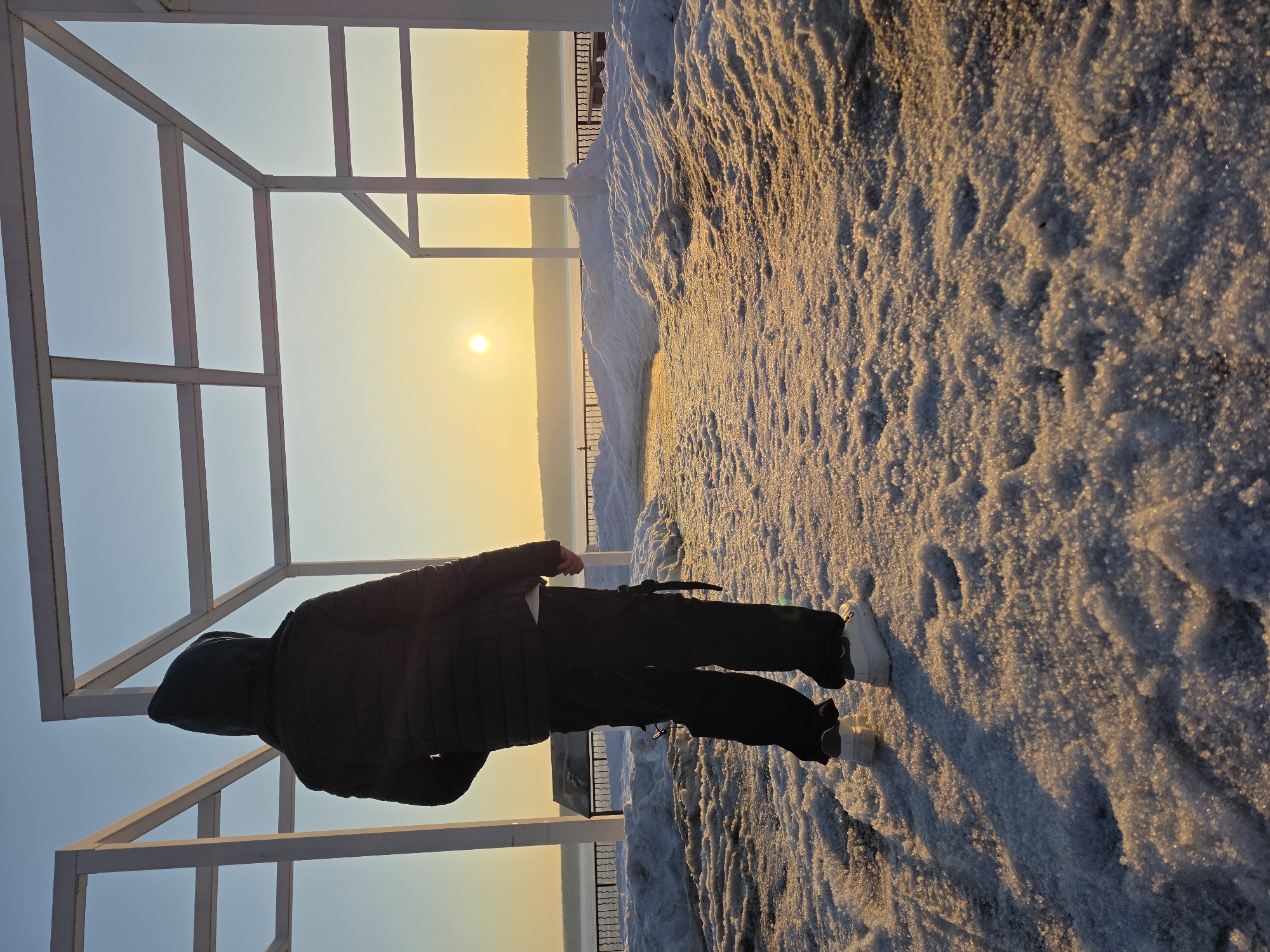 The scene depicts a solitary individual standing on a snow-covered promenade, likely the Embankment of the Kama River in Chaikovsky, Russia, during either sunrise or sunset. The person is observed from behind, facing towards a low sun positioned above a distant, tree-lined horizon.
The individual wears a dark, hooded puffer jacket, dark cargo-style pants with visible straps on the sides, and white sneakers. Their stance is still, with weight primarily on their left leg.
The immediate foreground consists of a textured layer of granular snow or ice, illuminated by the golden light of the sun, casting short shadows directly towards the viewer. The ground transitions from a clear, icy path directly in front of the individual to deeper drifts of snow on the sides.
Several white, open-frame rectangular architectural structures, possibly decorative arches or a pergola, frame the view, with some parts of these structures visible on the left and right sides of the image.
In the mid-ground, beyond the snow-covered promenade, a dark metal railing runs horizontally, indicating a boundary. Behind this railing, a wide body of water, likely the frozen or partially frozen Kama River, extends to the distant horizon. A dark, dense tree line forms the farthest visible element of the landscape, silhouetted against the bright sky. The sky transitions from a pale blue at the top to a bright golden-orange around the setting/rising sun.