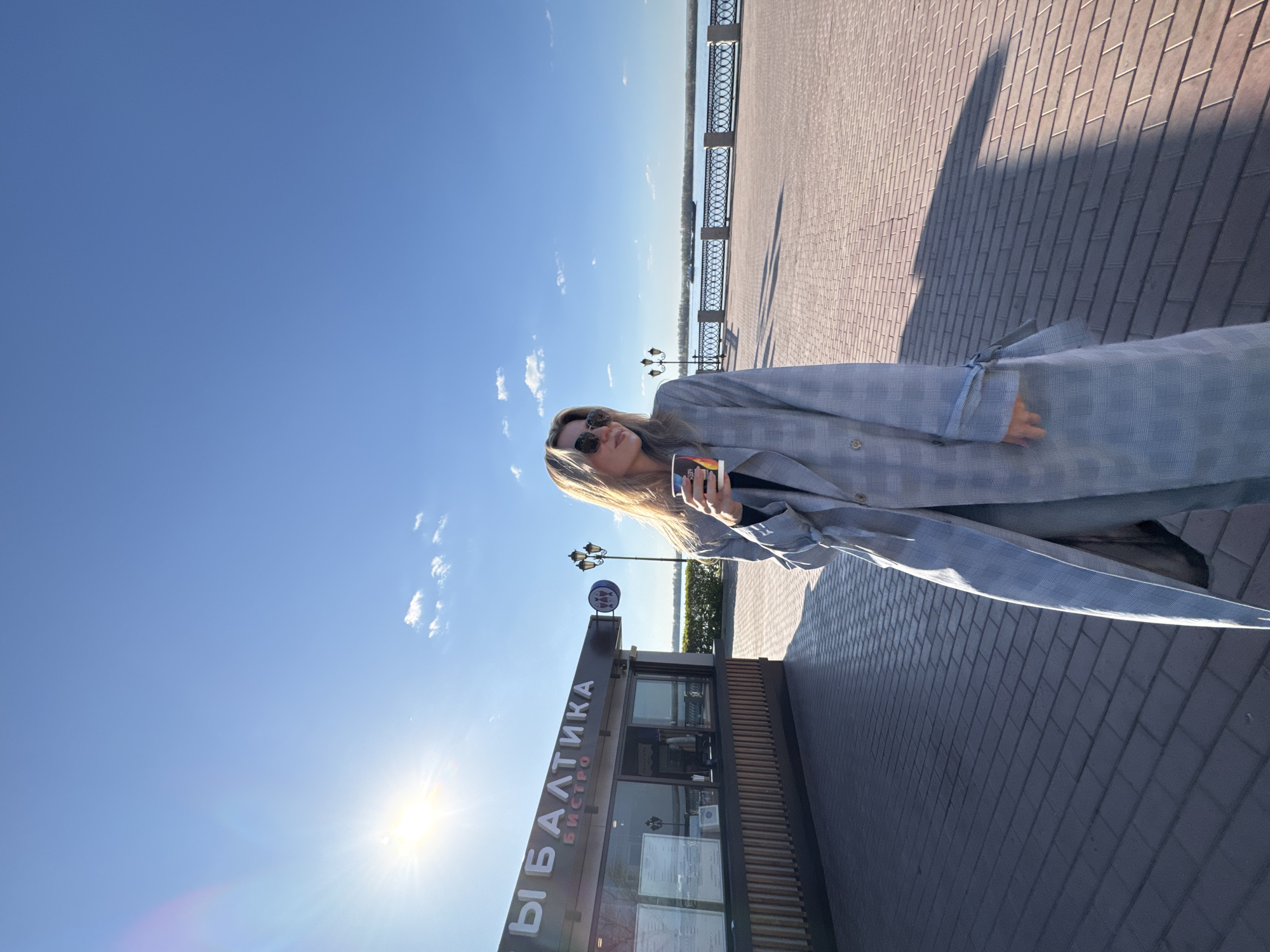 A woman wearing sunglasses and a plaid jacket is standing on a brick-paved area in Unknown, Unknown, holding a cup. Behind her is a building with the Cyrillic inscription "Ы БАЛТИКА БИСТРО". A river and a metal railing are visible in the background under a clear blue sky with a few clouds.