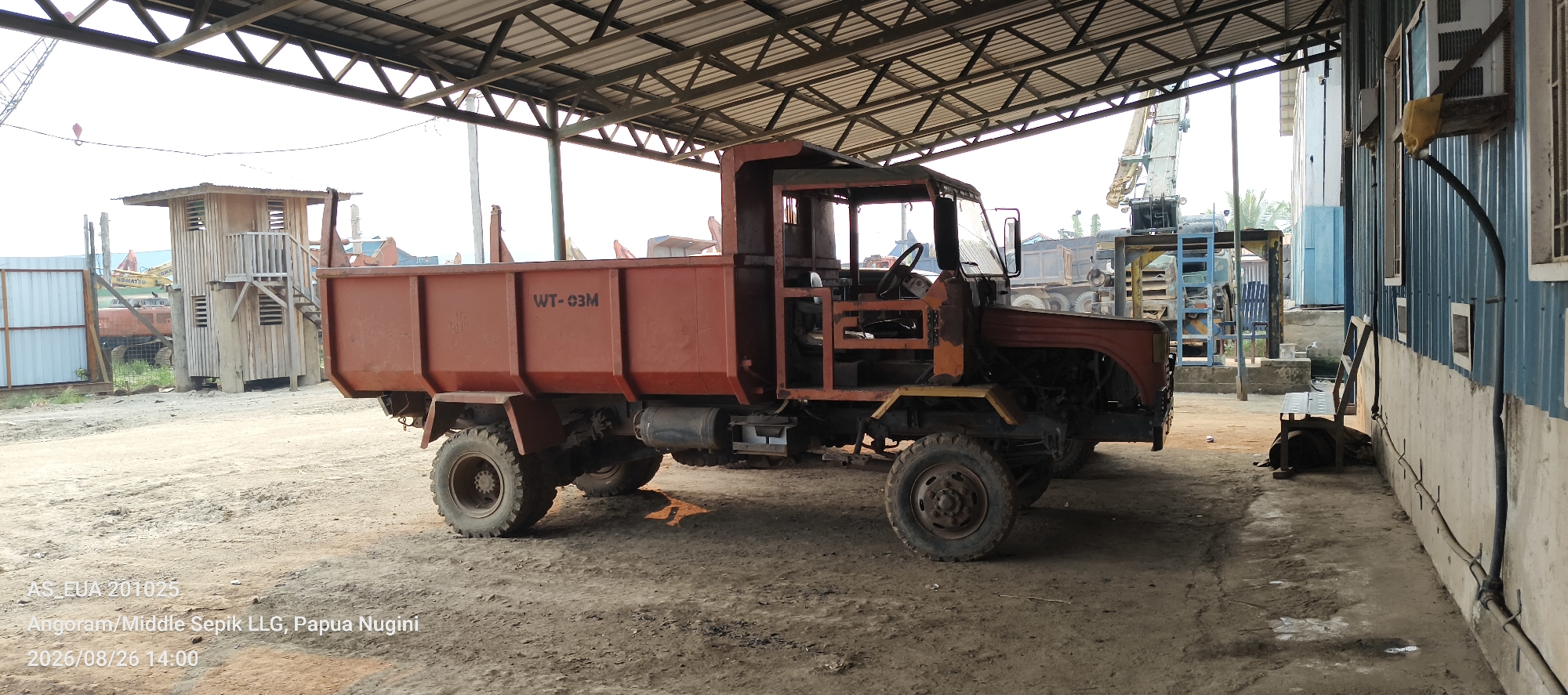 A rusty, orange dump truck is parked indoors under a metal roof structure. The truck is oriented diagonally, with its front facing right. The dump bed of the truck has "WT-03M" stenciled on its side. The driver's cab appears to be open and missing some parts. The ground is dirt and gravel. To the left of the truck is a small, wooden shack with a staircase leading to a raised platform. In the background, there are other vehicles and industrial equipment, including a large crane. To the right of the truck is a blue corrugated metal wall with windows and some electrical conduits. The scene is located in Angoram/Middle Sepik LLG, Papua New Guinea, as indicated by the timestamp and text overlay on the image.