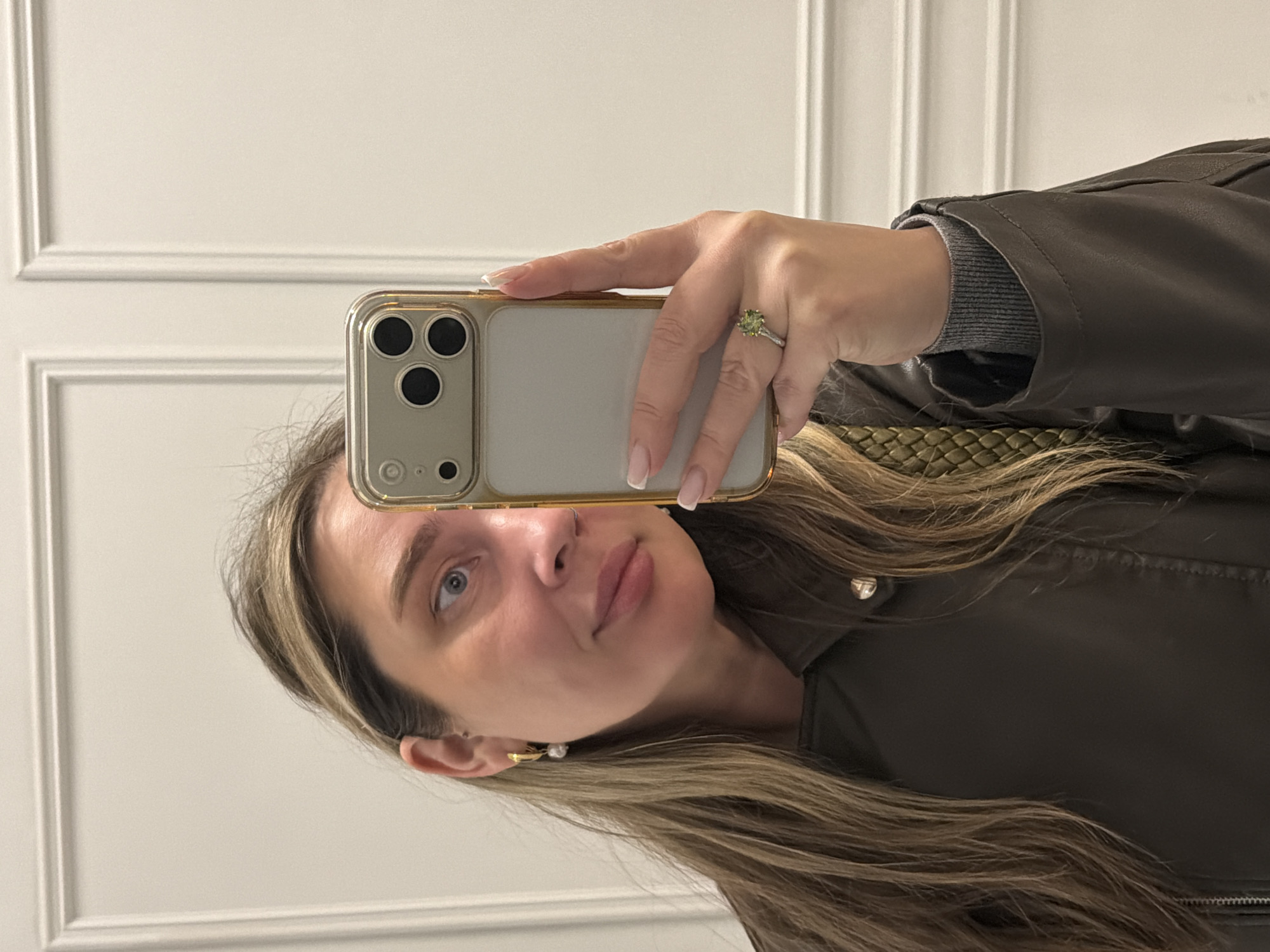 A woman is taking a selfie with her smartphone. She is wearing a brown leather jacket over a grey sweater. She has blonde hair and is wearing earrings and a ring. She is standing in front of a white wall with paneling. There is no indication of the location being Moscow, Russia.