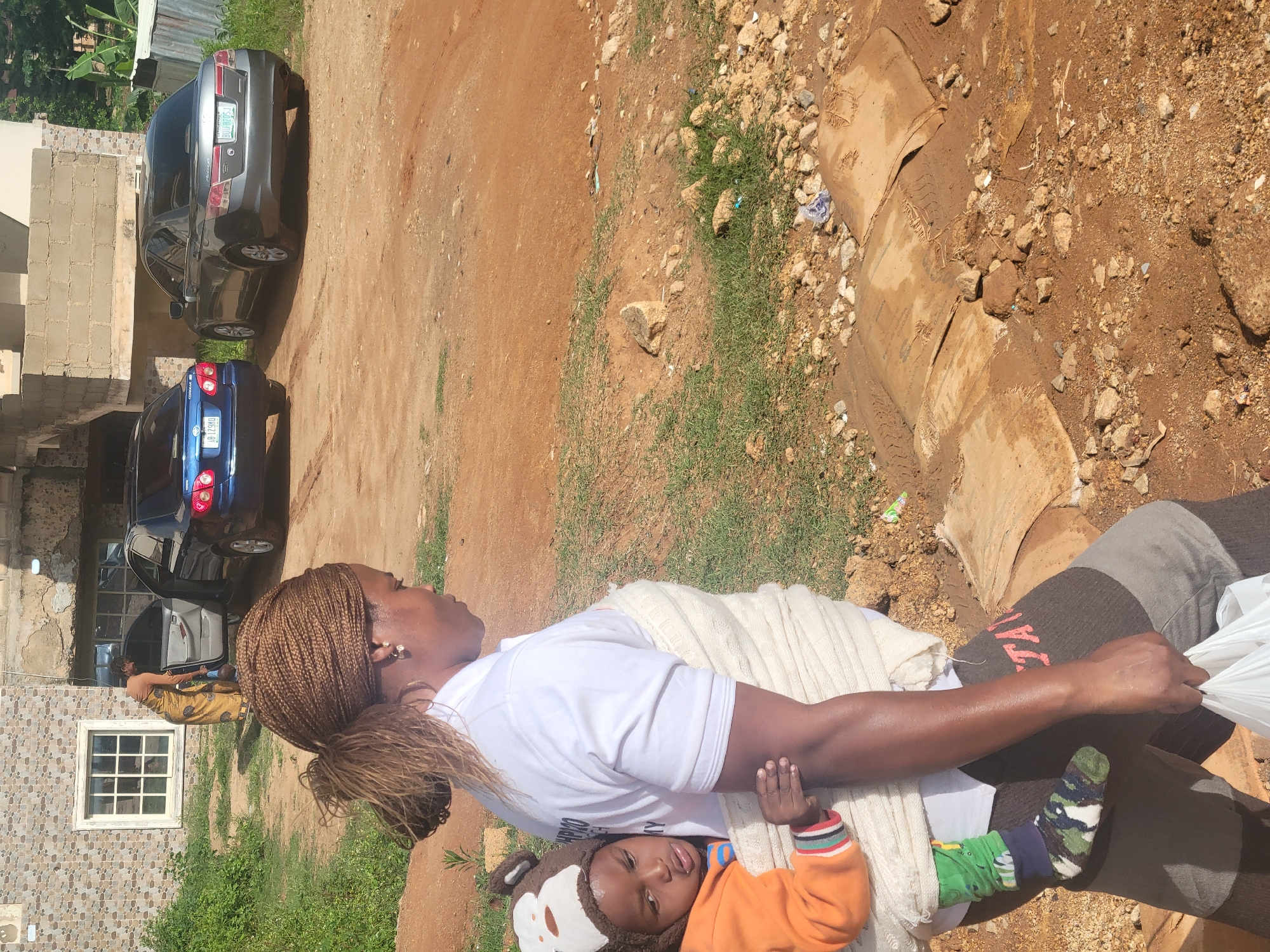A woman carries a child on her back, wrapped in a cream-colored blanket. The child wears an orange jacket and a brown hat with white trim. The woman has braided hair and wears a white t-shirt and brown patterned leggings. She holds a white plastic bag in her right hand. They are walking on a dirt path in front of a building with a tiled facade and a window. Behind them, two cars are parked on the dirt lot: a blue sedan and a gray sedan. The environment is somewhat unkempt, with sparse grass, rocks, and debris scattered on the ground. This scene is consistent with the outskirts of Jos, Nigeria, where unpaved roads and modest buildings are common.