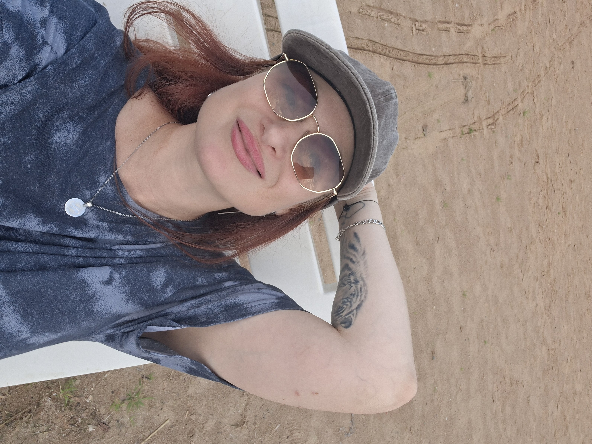A woman with red hair, wearing a grey cap and sunglasses, lies on a white lounger on a sandy surface. She is wearing a blue tie-dye t-shirt and a silver necklace. Her right arm is bent behind her head. The background is a sandy area with tire tracks and sparse vegetation. This appears to be a scene from the city of Myadzyel, Belarus.