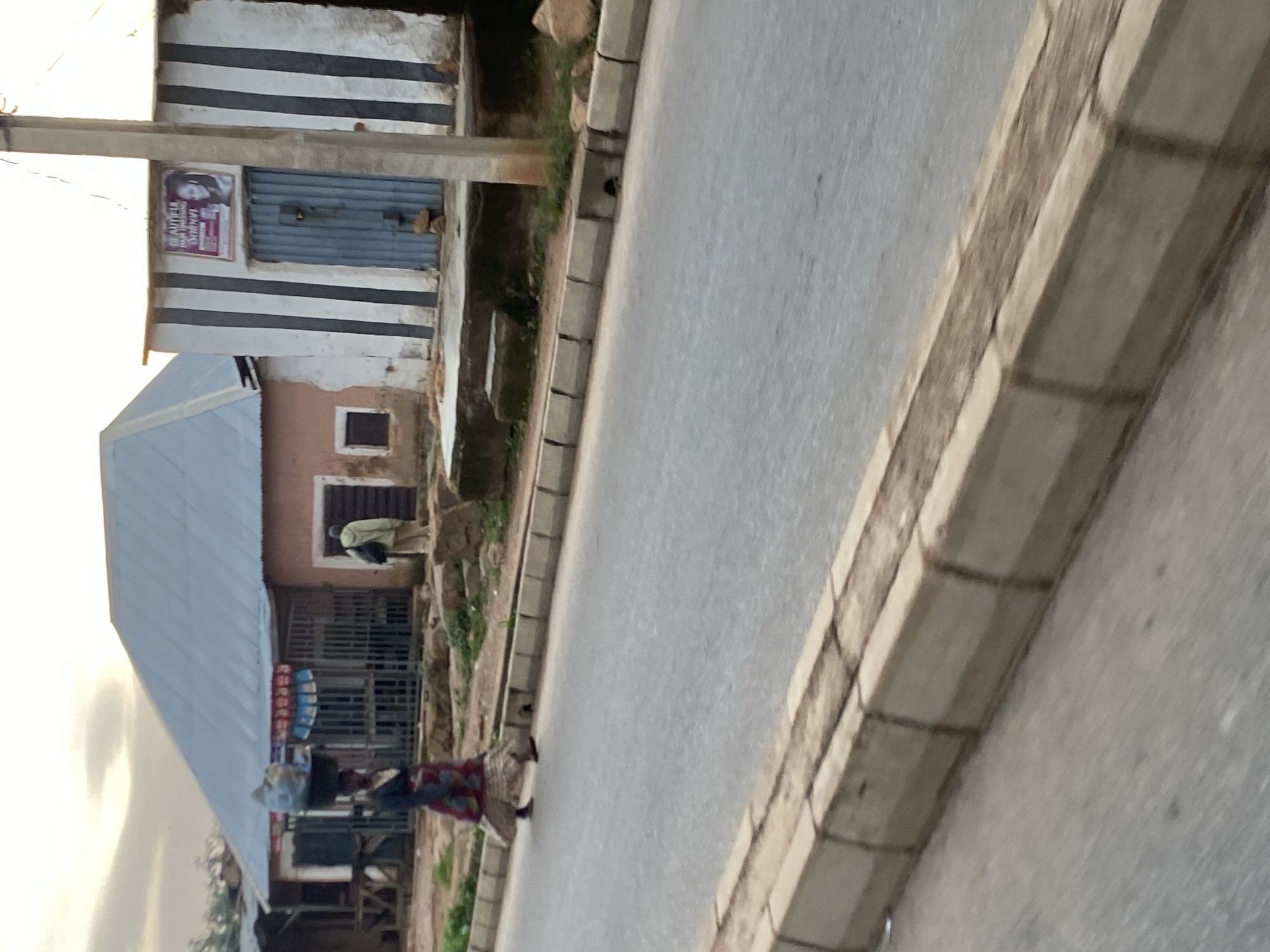 The image shows a street scene in Jos, Nigeria, with a woman carrying a load on her head walking on the side of the road. She is wearing a colorful skirt and a dark top. Behind her, a building with a silver roof and barred windows is visible. To the right, another building has vertical black and white stripes and a blue metal door. A sign advertising a "Beautiful Hair Dressing Salon" is attached to this building. The road is paved and bordered by a concrete curb. The sky is overcast. The overall impression is of a modest, possibly commercial, area in a city.
