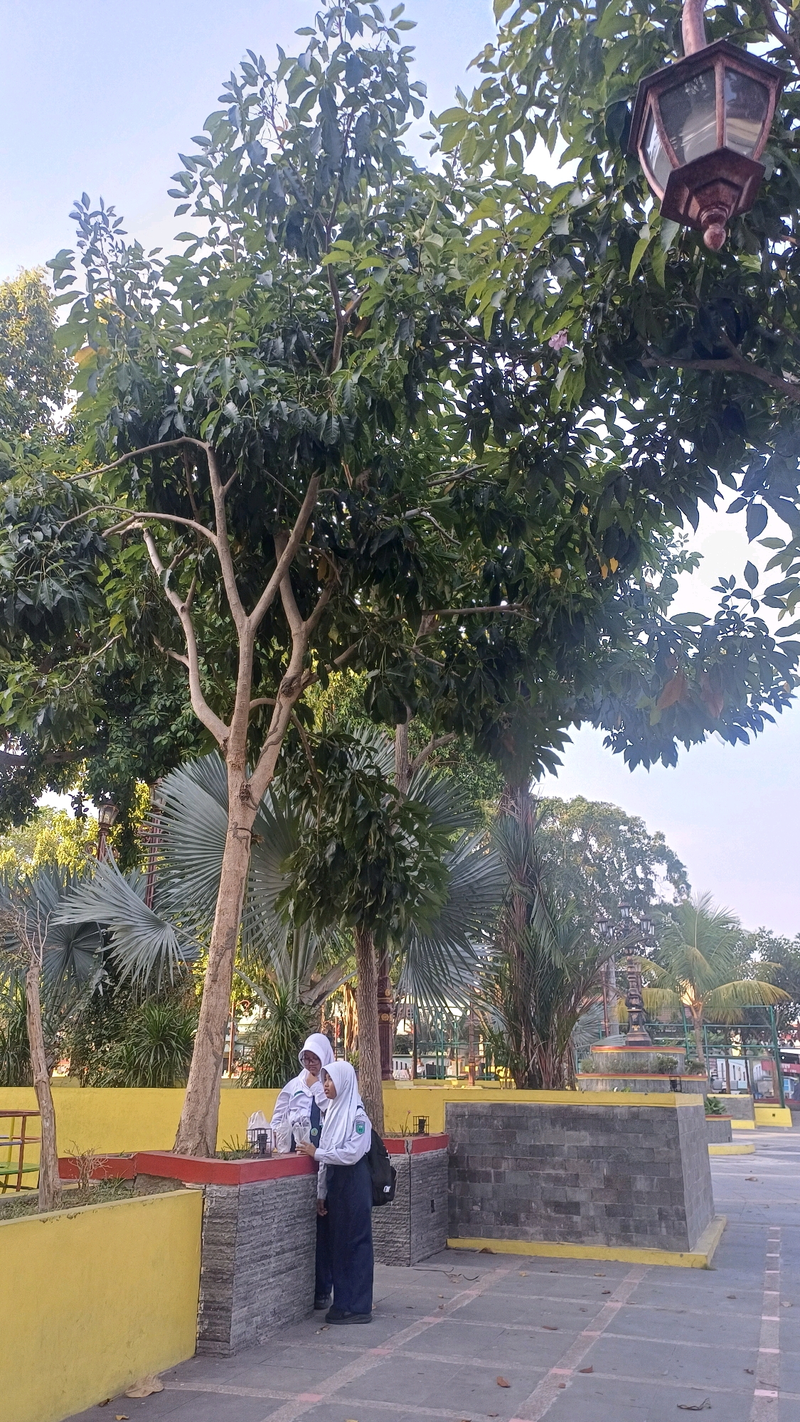 The image depicts an outdoor scene at the Klaten City Park in Klaten, Indonesia. In the foreground, two young people wearing school uniforms and hijabs are standing near a low, L-shaped wall. The person on the left is leaning on the wall, while the person on the right is looking towards the left. They appear to be engaged in a conversation. The wall is constructed with gray stone blocks and has a red border on top, with a bright yellow base. Behind them, lush green trees with dense foliage dominate the upper half of the image, with a street lamp visible through the branches on the right. In the background, more trees and park amenities like benches and decorative structures are visible. The ground is paved with square tiles. The sky is a light blue. The overall impression is of a peaceful park setting during daylight hours.