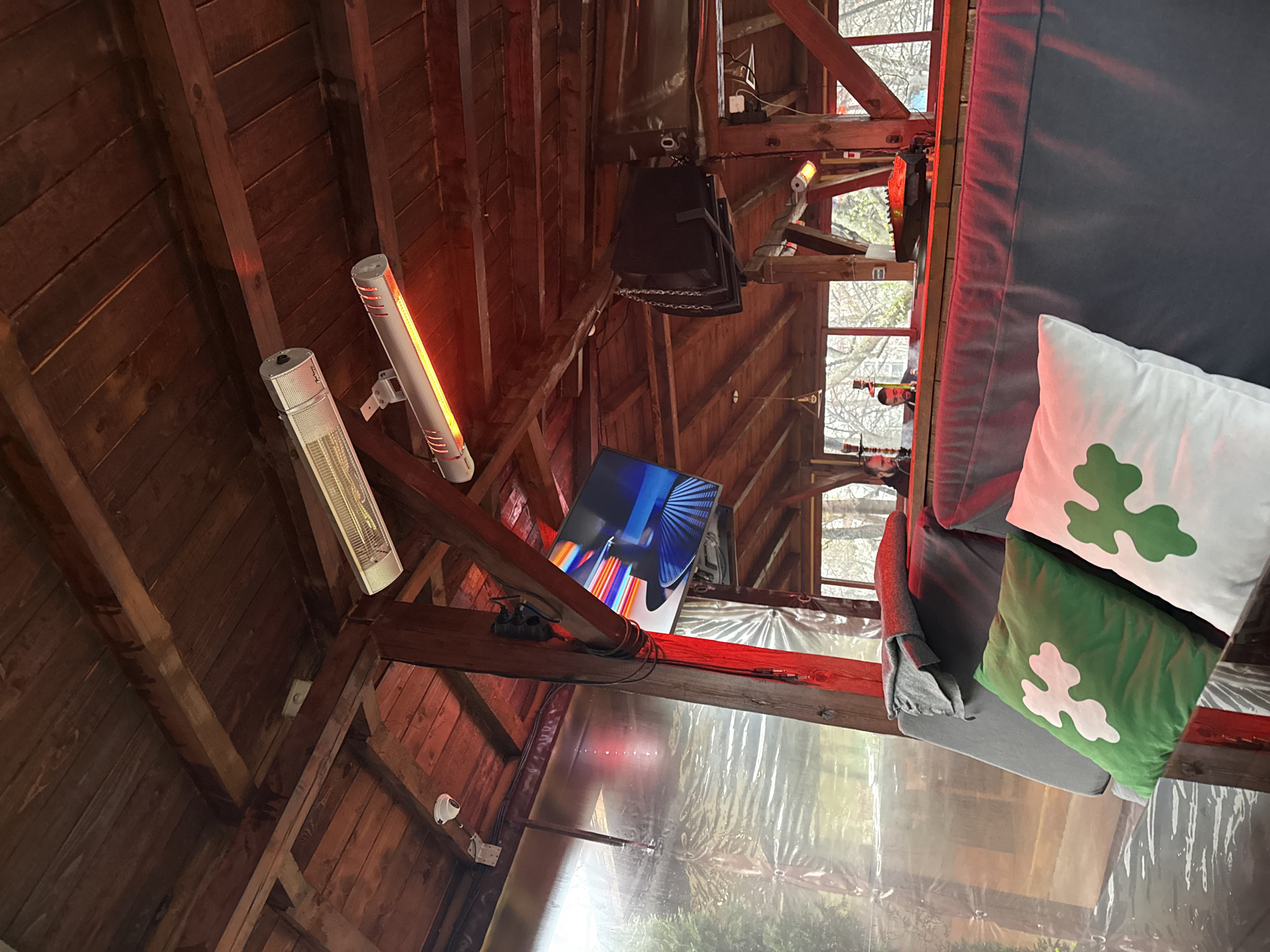 The image shows the interior of a wooden structure, possibly a shed or gazebo. The structure features exposed beams and a translucent plastic lining on the walls. Two electric heaters, one white and one with a glowing orange element, are mounted on the beams. A television screen displays colorful abstract imagery and is suspended from a beam. Also suspended from the ceiling is a dark-colored speaker box. A couch with pillows, including ones with a green shamrock design, sits in the foreground. The scene seems to be a customized or recreational space within a wooden structure. The city of Sofia, Bulgaria is not evident from the image.