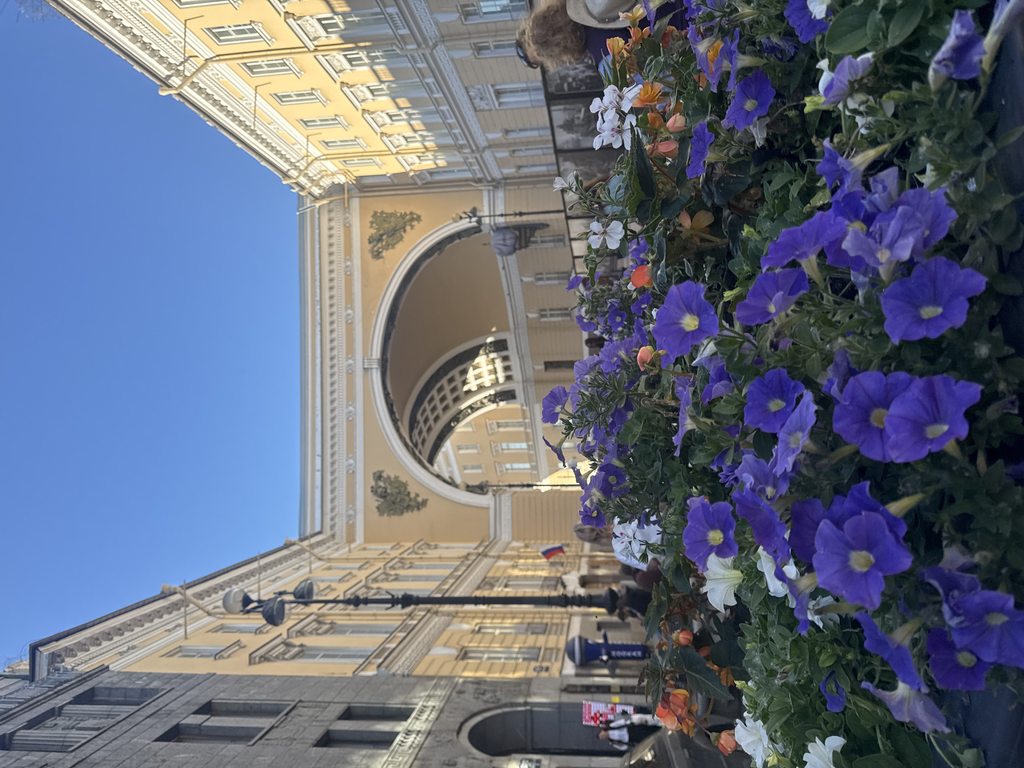 The image shows a foreground of vibrant purple and white petunias in a planter box, with a glimpse of orange flowers peeking through. Behind the flowers, a wide, arched entryway of a grand building is visible, with yellow facades on either side. The architecture suggests a historic urban setting. A lamppost stands to the left, and several people are walking in the background. A flag, possibly the Russian flag given the context of Saint Petersburg, is seen on a pole near the entryway. The sky is a clear, bright blue. This scene is likely in Palace Square in Saint Petersburg, Russia, specifically near the General Staff Building with its iconic arch.