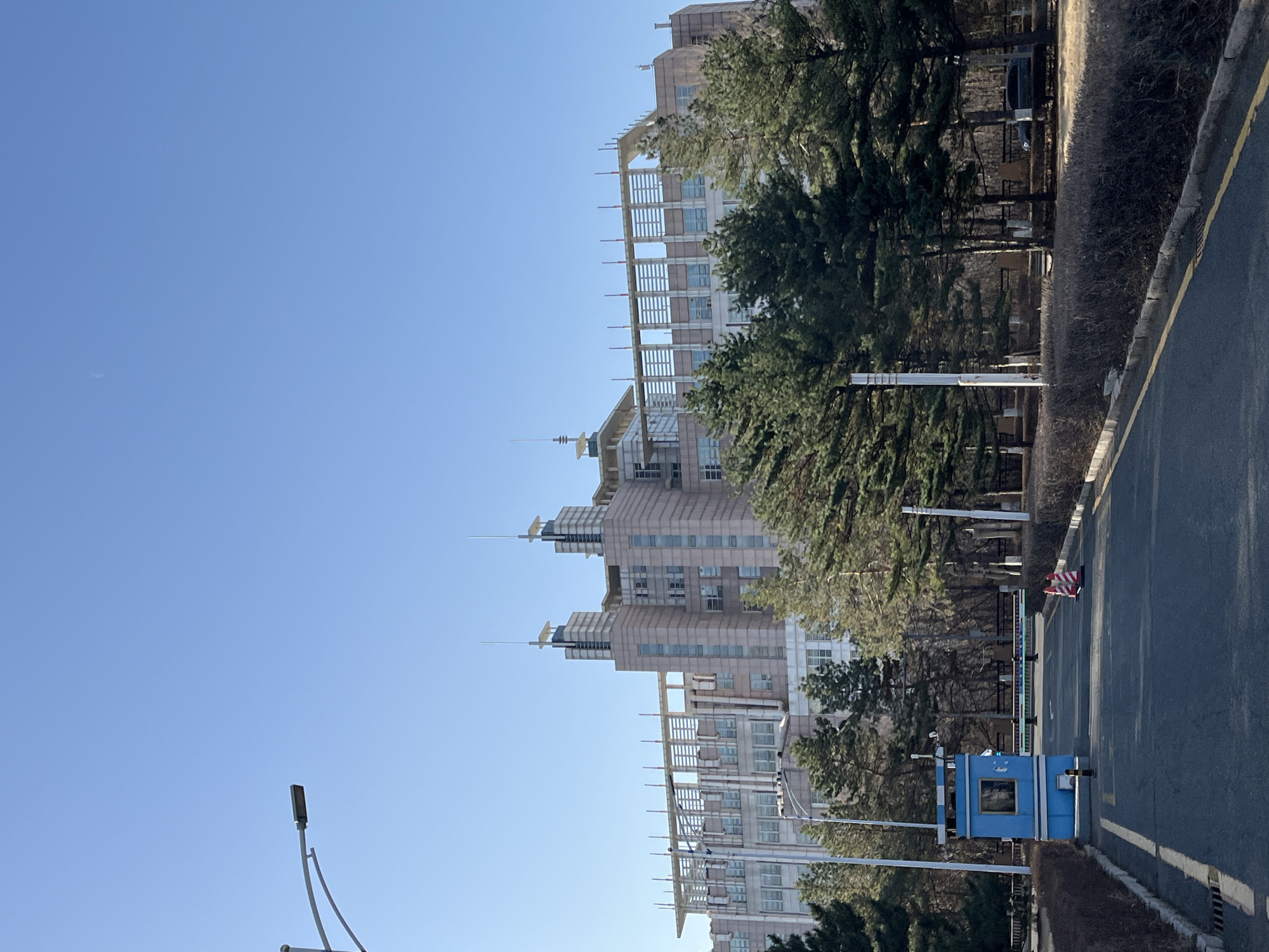 The image shows a view of buildings and street in Changchun City, China. The sky is clear blue, with a sliver of the moon visible. Multiple light-colored buildings with a layered design rise from the ground. Green trees partially obscure the buildings. A road stretches along the right side of the frame, marked with a yellow line. A small blue security booth stands on the road edge. Streetlights are positioned along the road. A fence separates the building area from the road.