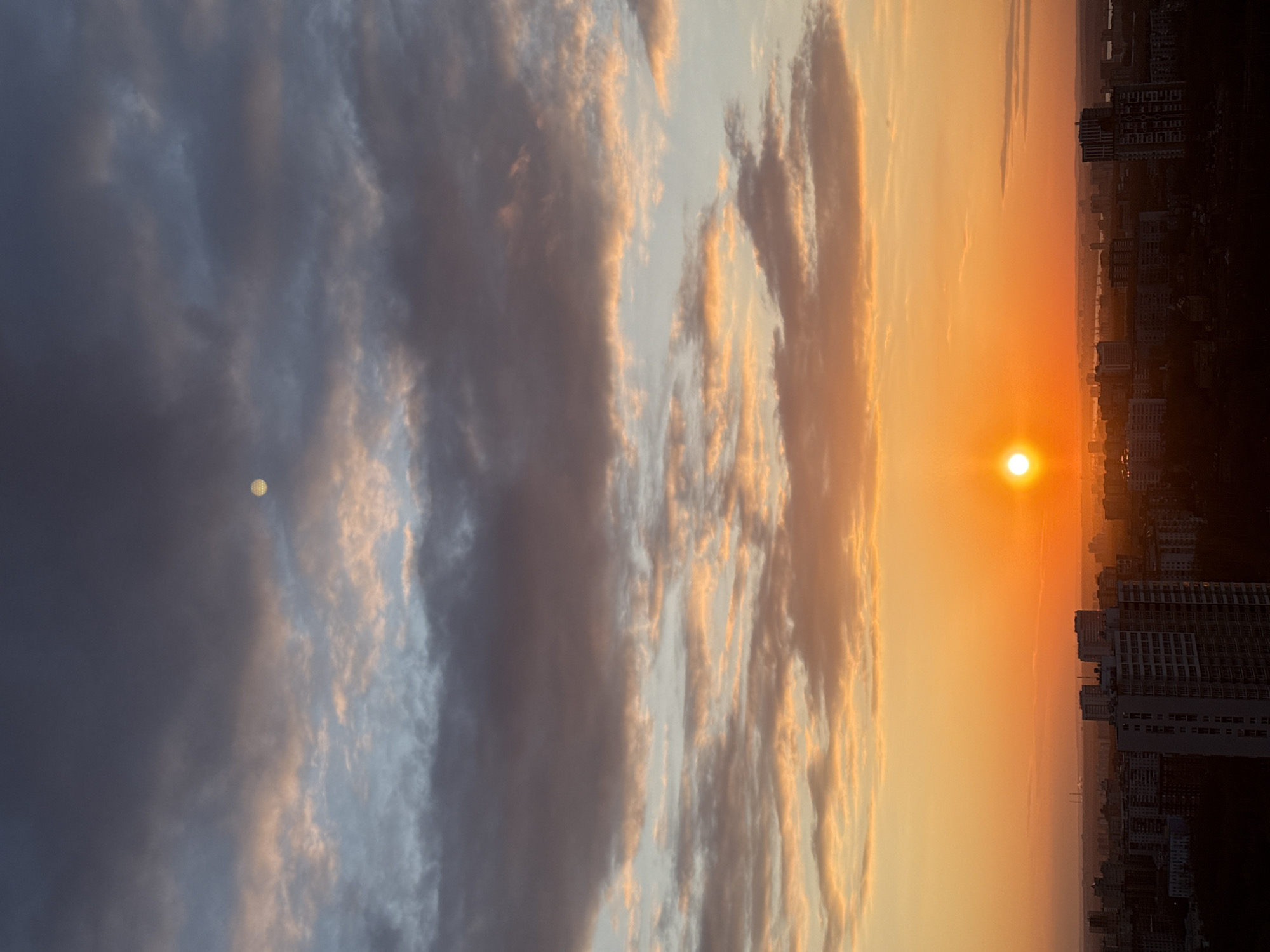 This rotated portrait photograph captures a dramatic sunset or sunrise over an urban skyline, likely in Perm, Russia. If viewed in its intended landscape orientation, the sky occupies the upper portion, and the city forms the horizon line below.
The sky is a vibrant canvas of contrasting light and shadow. On the left side, dark, heavy clouds dominate, appearing in shades of grey and deep blue, tinged with subtle pink and orange hues from the distant light. Within this darker cloud mass, a small, circular, white object with a discernible grid-like or cellular texture is visible, suggesting it is the moon.
The right side of the sky transitions into a brilliant expanse of glowing orange and yellow, indicating the presence of the low-lying sun. The sun itself appears as a bright, circular, intense yellow-orange orb, positioned towards the right of the frame, just above the distant horizon. Streaks of lighter, wispy clouds are illuminated by its light, creating warm tones against the cooler, darker clouds further away.
Beneath the radiant sky, the silhouette of a city stretches across the horizon. Tall, modern high-rise buildings are clearly visible on the right, their dark forms sharply outlined against the fiery backdrop. The urban sprawl extends into a hazy distance, suggesting a large metropolitan area. The overall scene evokes a sense of peaceful grandeur at either dawn or dusk. There is no visible text in the image.