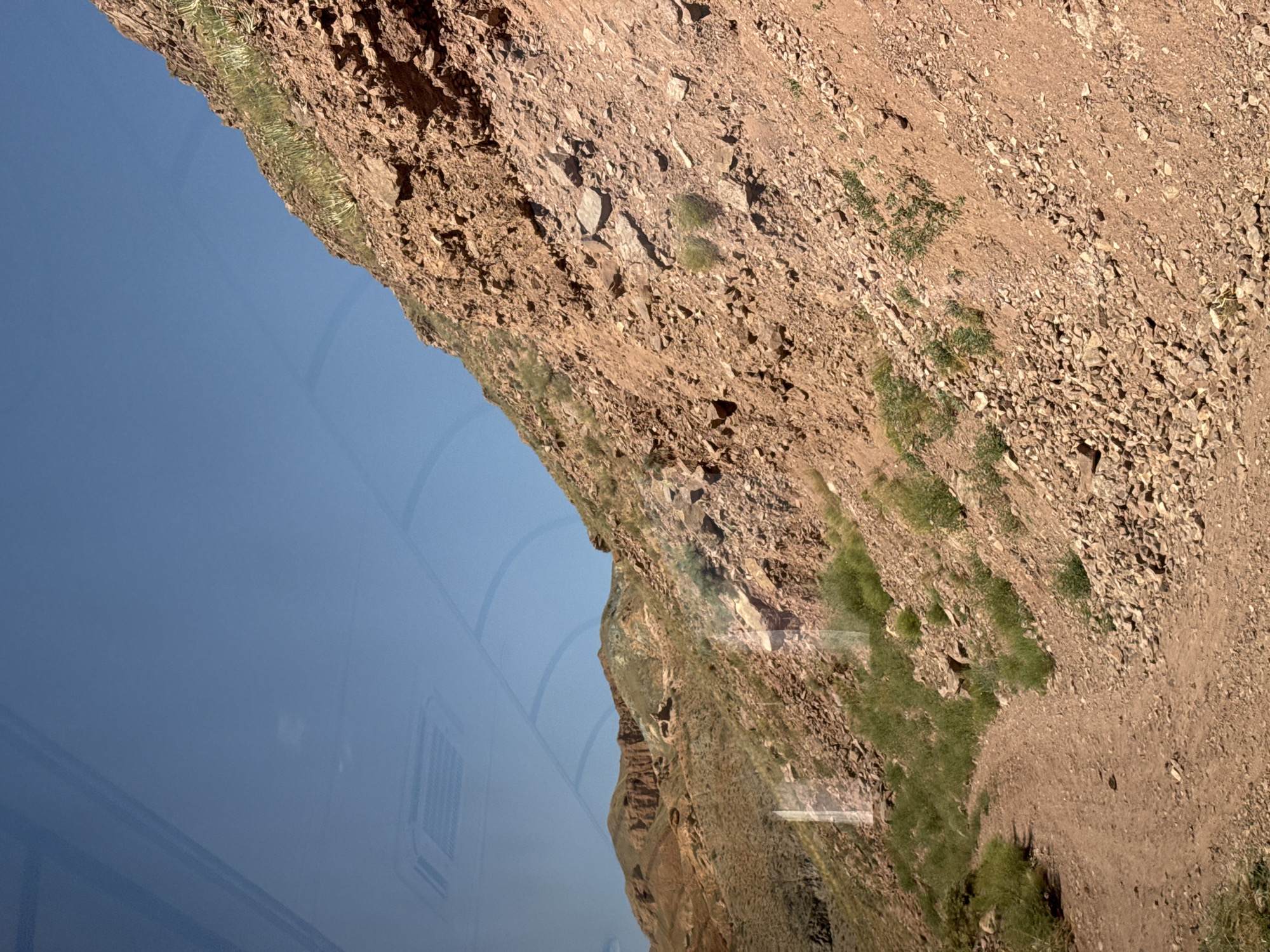 The image shows a dirt road winding up a rocky, reddish-brown hillside under a clear blue sky. Sparse green vegetation, appearing as tufts of grass and some larger plants, grows along the road and on the slopes. In the distance, a range of similar hills is visible, with more eroded formations. The overall environment suggests a dry, arid landscape. There are some reflections on the surface, implying the photo was taken from inside a vehicle or through a window. No people or specific landmarks are visible in the scene, nor is there any indication of the city of Krasnyy Most, Kyrgyzstan, within the visual information presented.