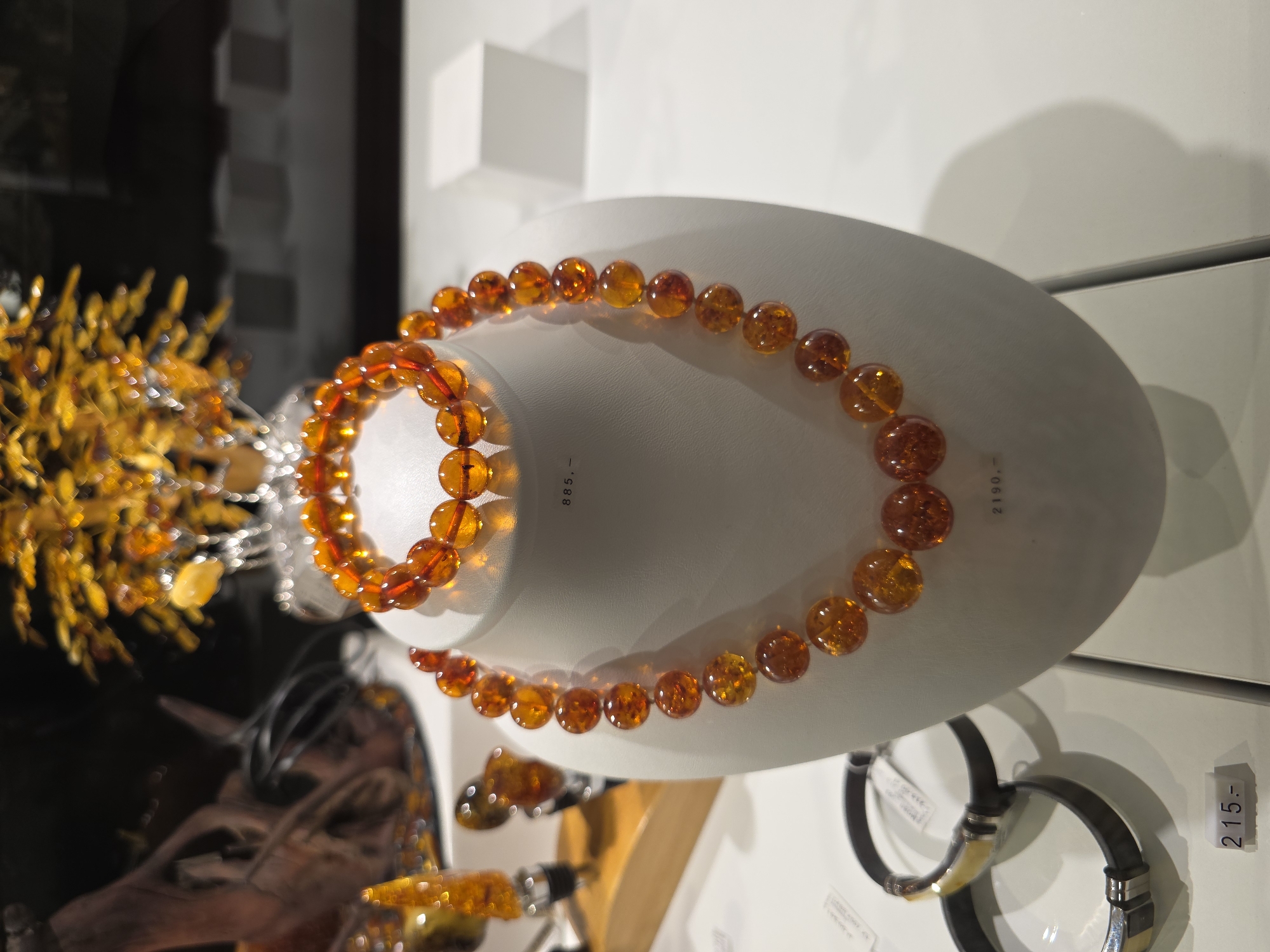 The image captures a display of amber jewelry. A beaded amber necklace and bracelet are draped over a white, oval display stand. The amber beads are a rich orange-brown color. To the left, there is another amber display, possibly a decorative tree or bouquet made of amber pieces and silver wire. In the background, various other pieces of jewelry can be seen on display. Price tags are visible beside some of the items. The scene appears to be set in a jewelry store or museum exhibit. Based on the artifacts being presented (amber jewelry), the city of Warsaw, Poland is a plausible location.