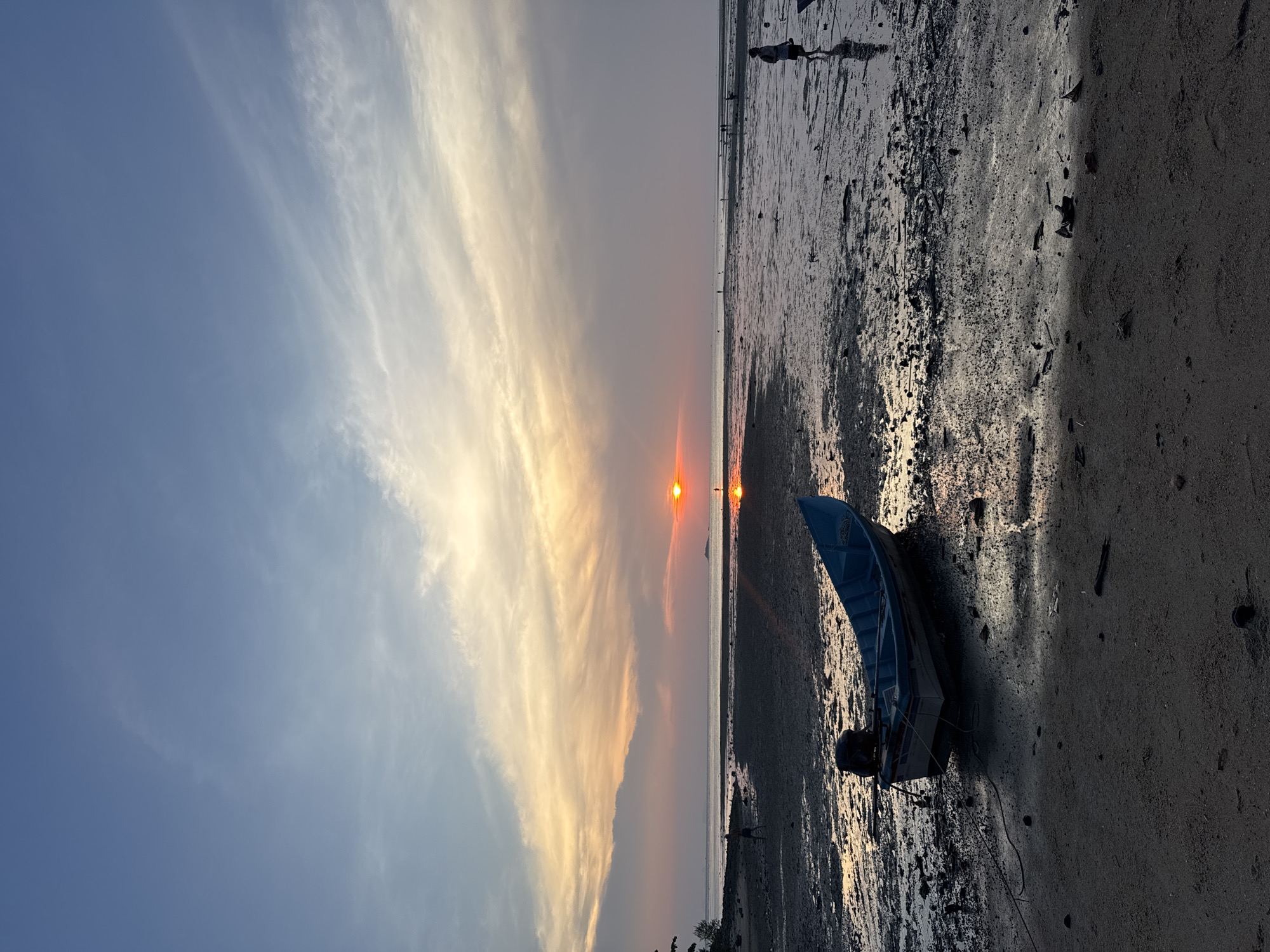 This media file captures a serene coastal landscape at either sunrise or sunset in Baan Wok Tum, Thailand. The horizon line stretches across the image, dividing the sky from the expansive beach and water.
The sky, occupying the upper portion, transitions from a clear, soft blue on the left to warm golden and orange tones towards the right, where the sun is low on the horizon. Long, wispy clouds streak across the sky, illuminated by the sun's soft glow.
The sun, a bright orange-red orb, is visible just above the distant horizon, casting a shimmering golden path of light across the calm water and wet sand. The lower half of the image features wide, dark mudflats or wet sand, reflecting the sky's colors and the sun's brilliance. The texture of the ground appears damp and uneven, characteristic of a tidal area.
In the foreground on the right, a small, blue and white boat is beached on the wet sand, seemingly left there during low tide. Its hull is angled, and some lines or ropes are visible near it. Several distant human figures are scattered along the horizon, appearing as silhouettes against the radiant light, walking or wading through the shallow water or mud. One figure is more clearly visible closer to the foreground on the right, near the boat.
The scene conveys a peaceful atmosphere, highlighting the natural beauty of the coastline during the golden hour. There is no visible text in the image.