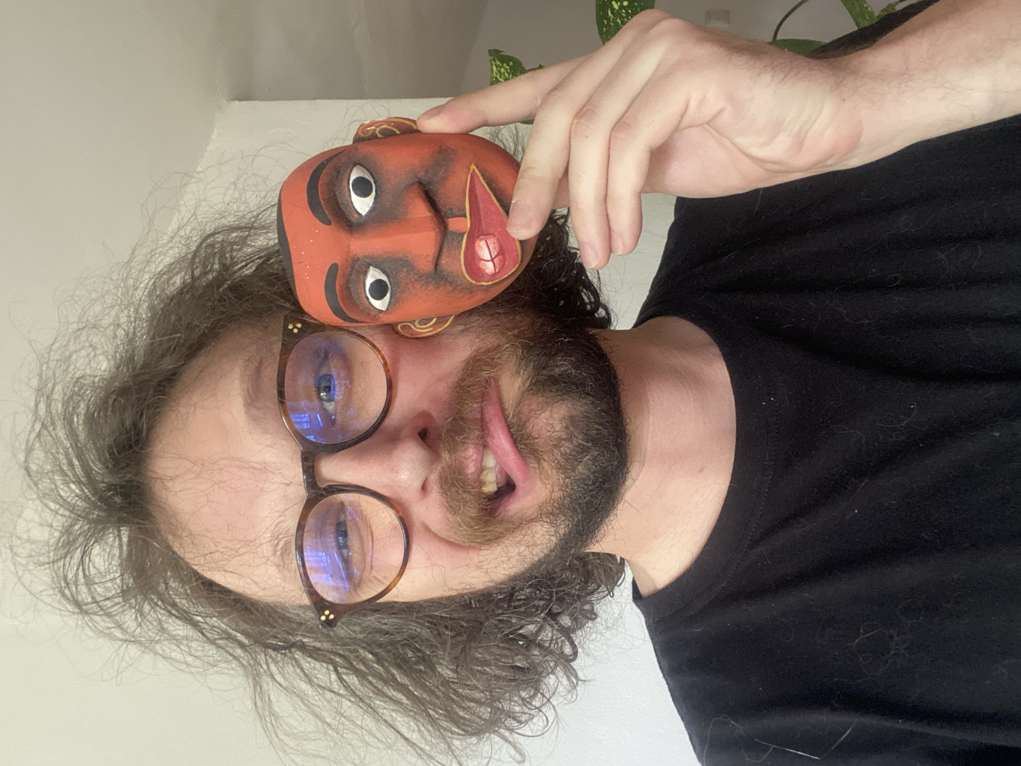 A selfie shows a man with long, curly brown hair and a beard, wearing round glasses, holding a red mask next to his face. The mask has painted white eyes and a decorative teardrop shape on its cheek. He is wearing a black t-shirt. The background is a white wall and a glimpse of a green plant. There is no indication in this image that it was taken in Rusanovo, Russia.