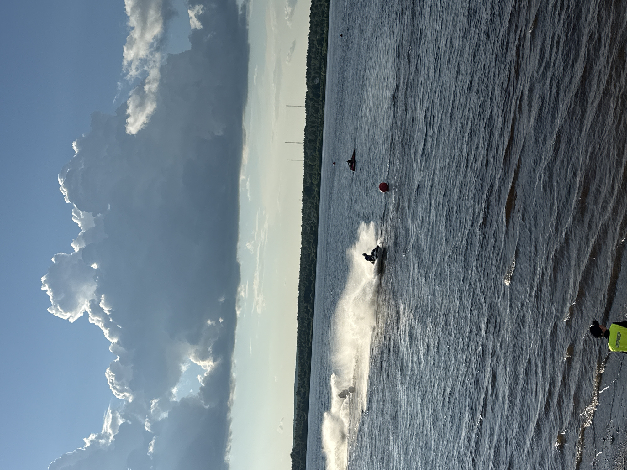 The image depicts a wide, dark blue body of water, likely a river or lake, under a dramatic sky filled with large, white cumulus clouds. The sun is setting or rising, casting a warm light on the scene. Several jet skis are visible on the water, with two in the foreground actively creating large splashes as they speed. One jet ski in the mid-ground is stationary, and another further in the distance. Red buoys are scattered in the water, suggesting a course or designated area. The far bank of the water is lined with a dense, dark green forest, and a few tall, slender structures, possibly communication towers, are visible against the horizon. In the bottom left corner, a person wearing a bright yellow vest with Cyrillic lettering and a black cap stands on a dark, wet shoreline. This scene is located on the Kama River in Perm, Russia.