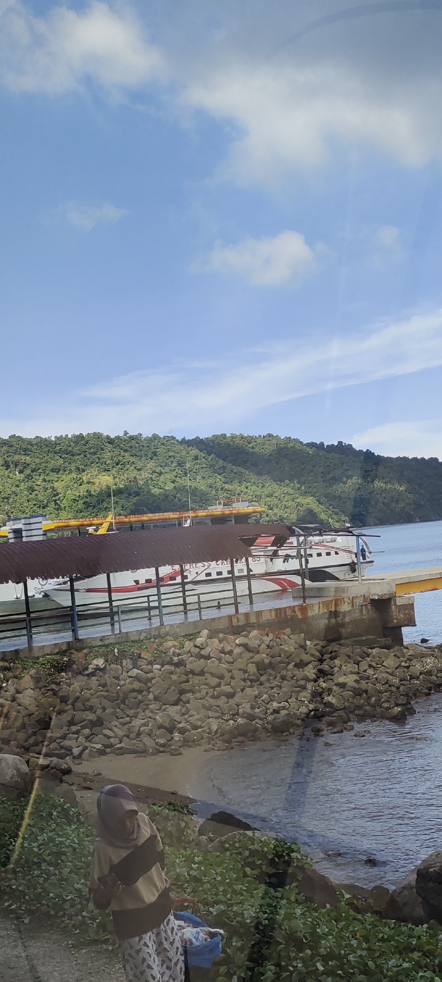 The image depicts a waterfront scene in Sabang, Indonesia, under a partly cloudy blue sky. In the midground, a ferry, identified by "EXPRESS CAR" lettering on its side, is docked at a pier. The ferry is white with red accents. The pier itself is constructed of concrete and is weathered, with a rusted metal roof supported by posts extending over the water. Rocks and vegetation are visible along the edge of the shore in the foreground. A person wearing a headscarf and a striped shirt is standing near the water, carrying a blue bag. The background features a densely vegetated, green hillside overlooking the water. The scene suggests a functional port or transportation hub.