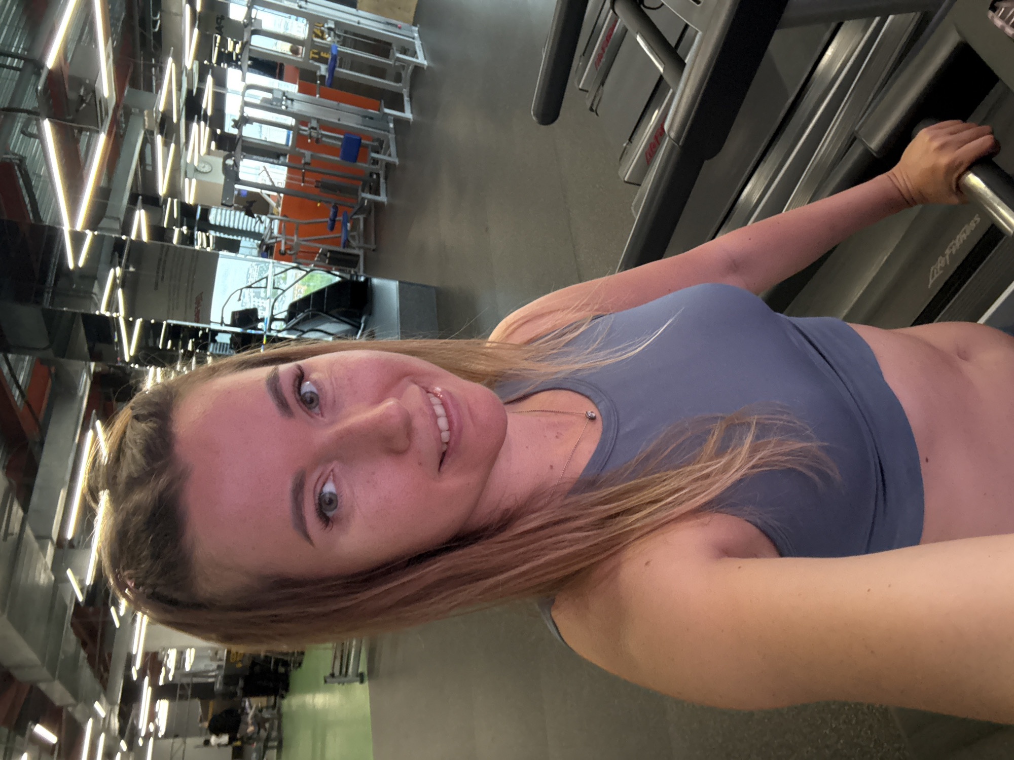 A person, presumably female, is at a gym in Unknown, Unknown. She is on a treadmill and holding on to the front support bar. She is wearing a gray sports top. The gym is well lit with modern lighting. There are weight lifting machines in the background.