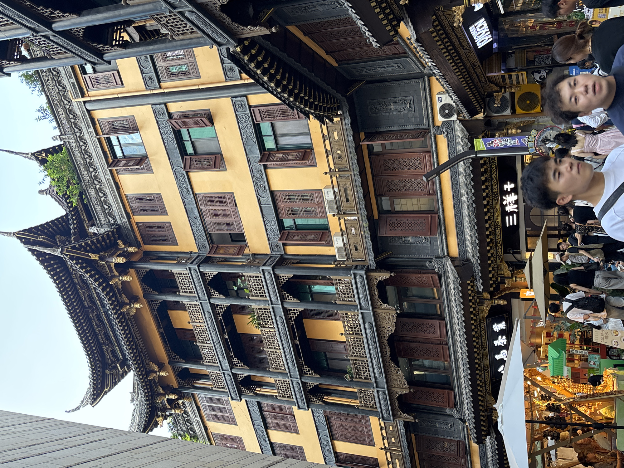 This image shows a street scene in Yuzhong District, China, featuring traditional Chinese architecture with a modern twist. A multi-story building with yellow walls and dark wood accents dominates the frame. The building has intricately carved wooden balconies and windows, with some shutters open. The roofline features a classic curved design with ornate details. To the left of this building, a modern grey structure is visible, suggesting a mix of old and new in the urban landscape.
In the foreground, several people are walking along the street. Some are casually dressed, while others appear to be tourists. A market stall with white and beige umbrellas is set up, displaying various items, including what appear to be dried goods and decorative items. Signs in Chinese characters are visible on the buildings, indicating businesses or establishments. One prominent sign above the market stall reads "三祥", and another neon sign in English reads "1919CN". The presence of air conditioning units on the buildings and the modern clothing of the people indicate that this is a contemporary urban environment despite the traditional architectural style. The overall impression is a bustling street scene where cultural heritage is integrated with modern city life.