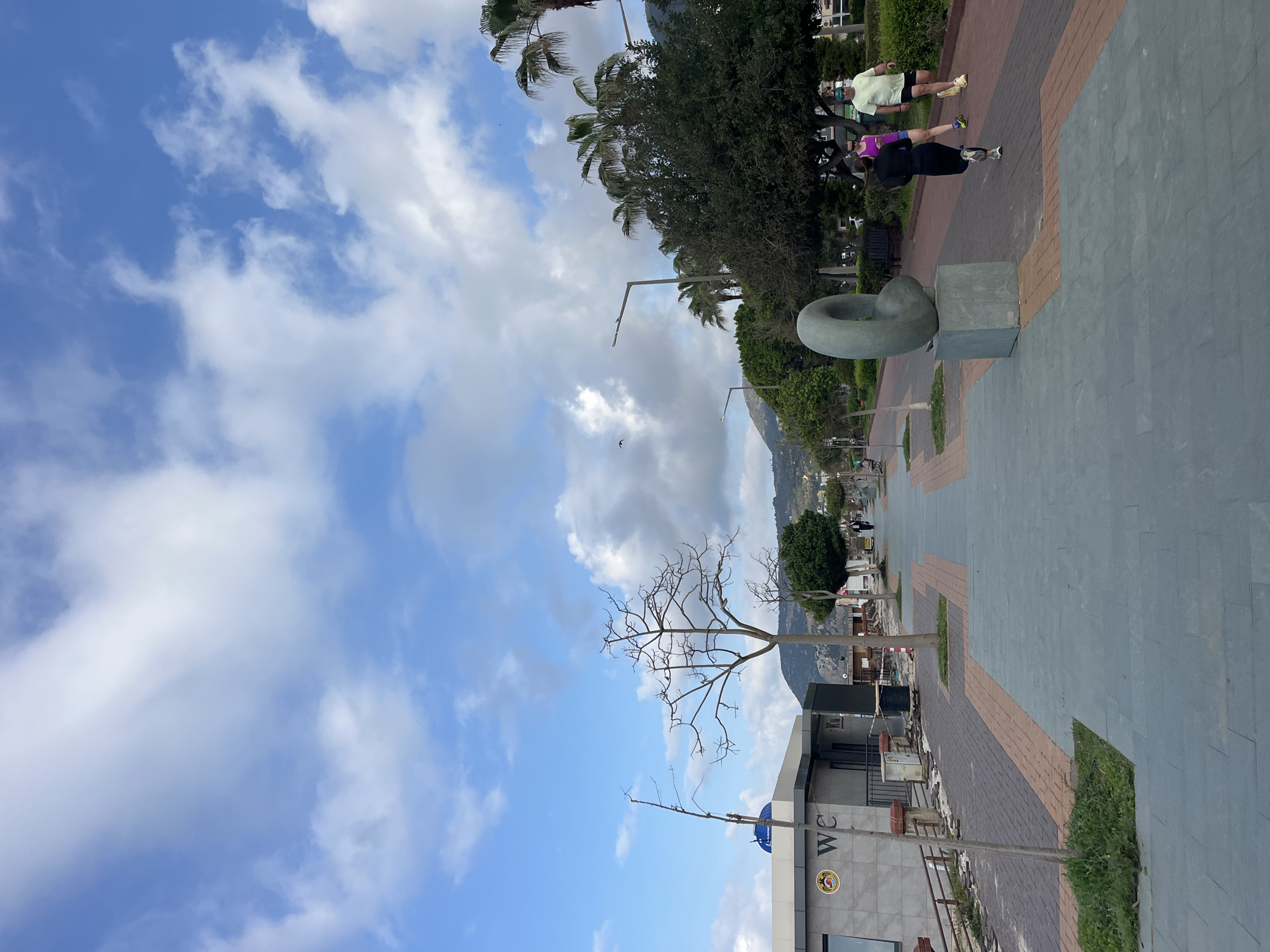 The image depicts a waterfront promenade in Alanya, Turkey. A bright blue sky filled with puffy white clouds is above. The promenade is paved with grey and brown brick and stone. There's a modern sculpture of a twisted ring on a concrete plinth. Two people are jogging in the distance, a woman in a purple top and a man in a white shirt. Palm trees and lush greenery are visible alongside the promenade. In the background is a distant mountain range. A bare tree is prominently displayed with the "WC" public restroom building in the bottom portion of the frame.