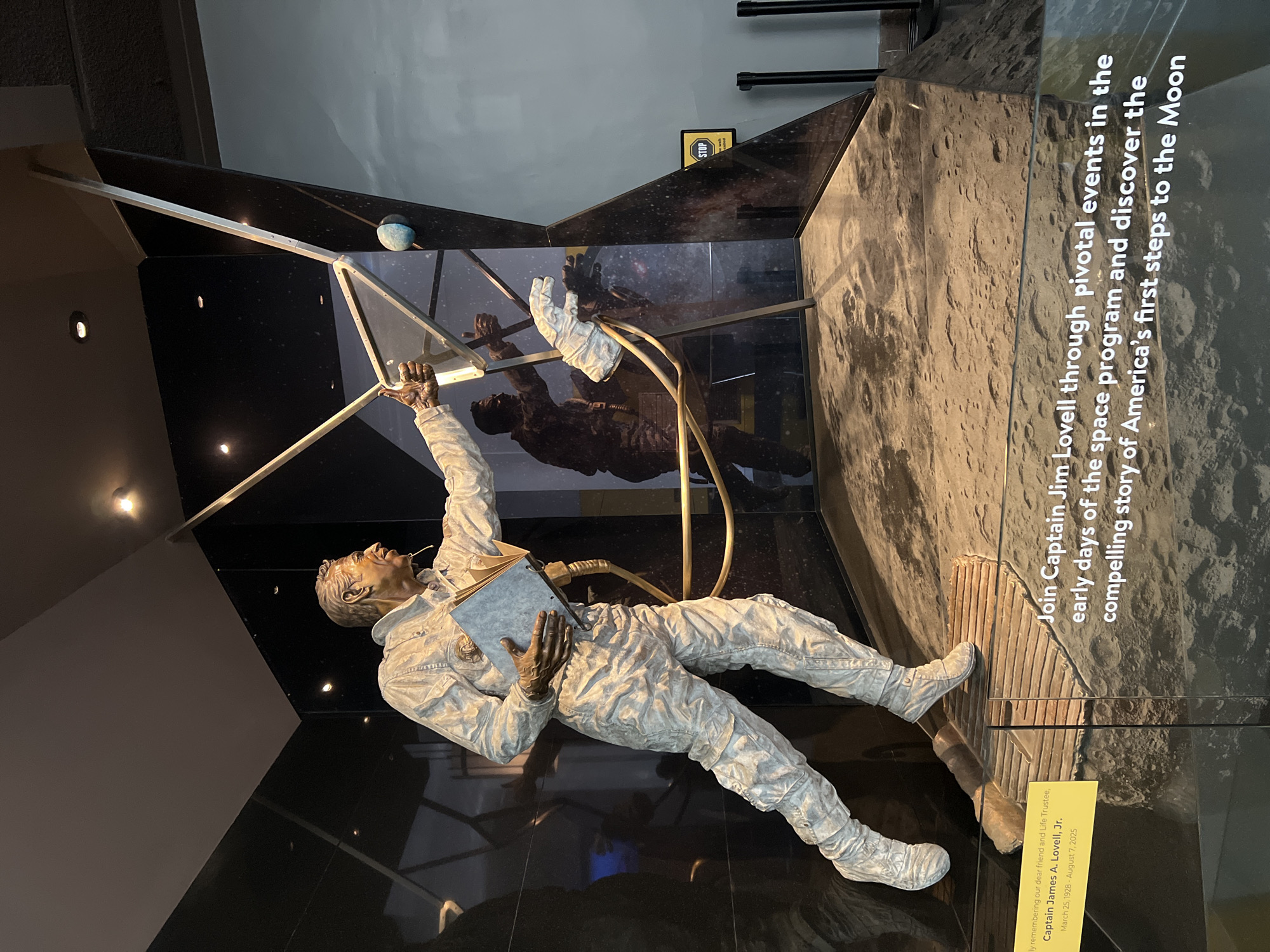 The image shows a statue of Captain James A. Lovell, Jr. dressed in a space suit holding a book, situated within a museum-like display. The display includes reflective surfaces, a miniature blue planet, and a plaque dedicating the exhibit to Lovell. The statue appears to be reaching toward a geometric structure. The exhibit aims to illustrate Lovell's role in the early days of the space program and America's first steps to the Moon. The exhibit is located in the city of Chicago, United States of America.
